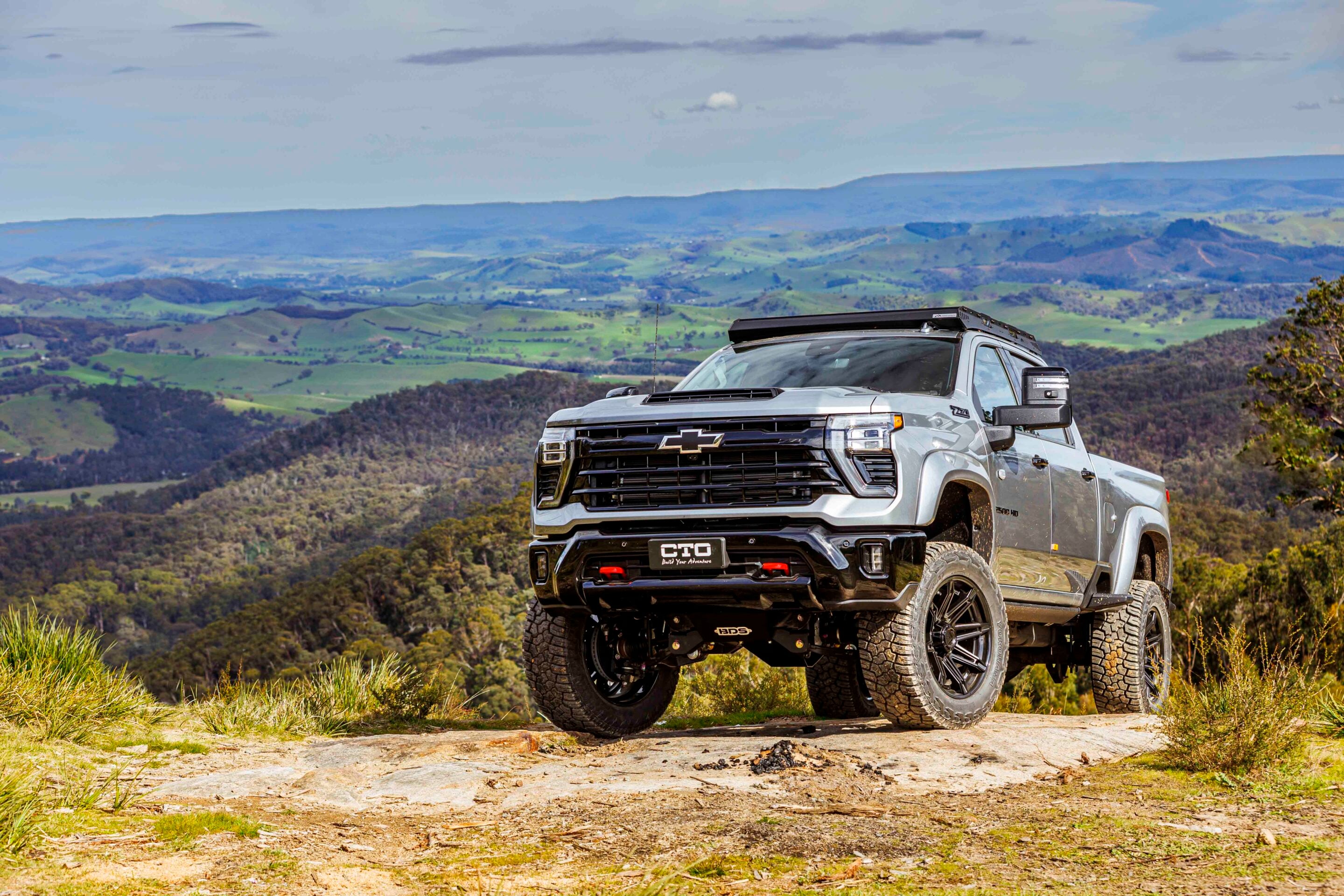 Silver Chevrolet pickup parked on a dirt trail with green rolling hills and blue sky in the background.