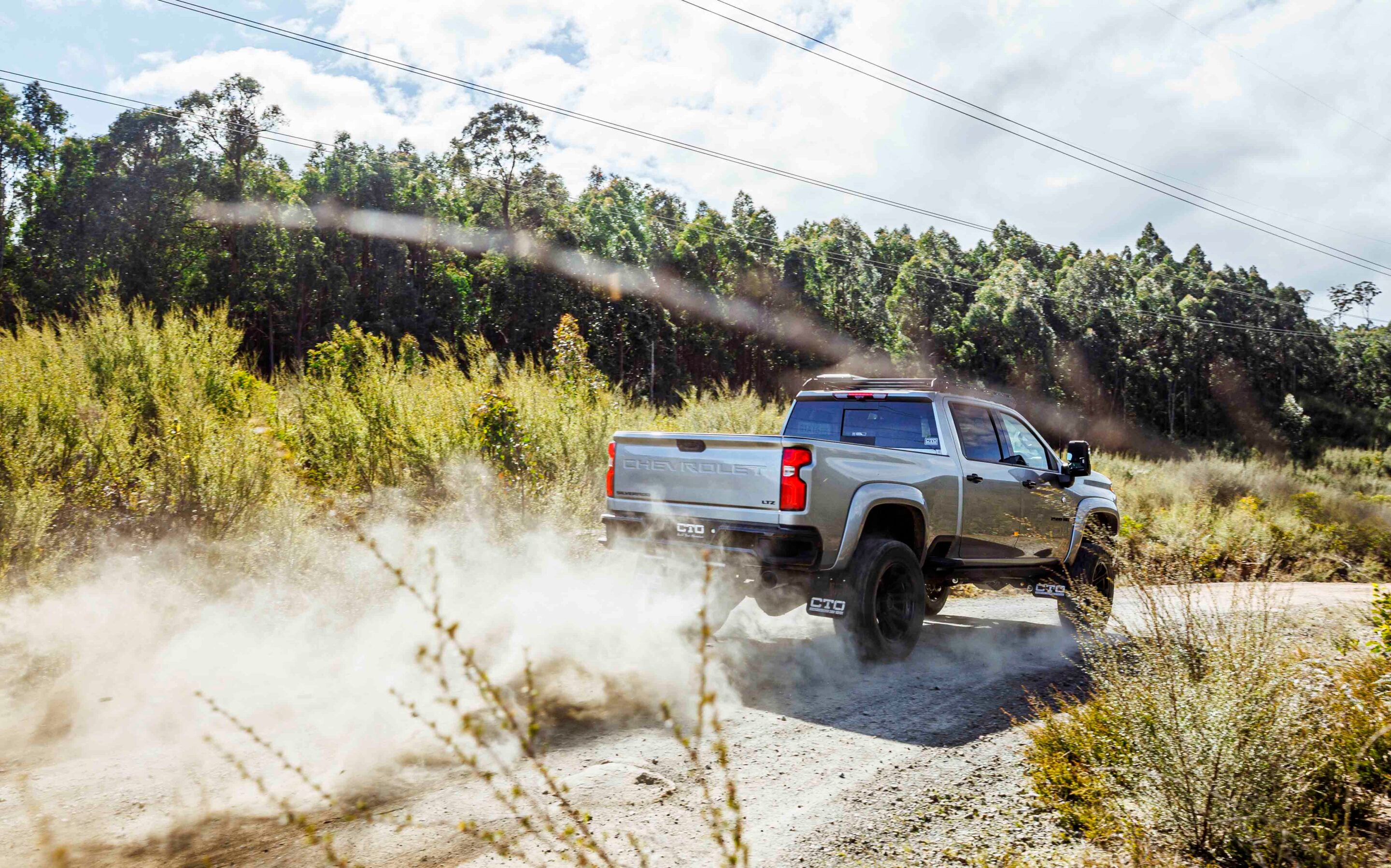Silver Chevrolet pickup driving off-road, kicking up dust on a dirt path with tall grasses and a forested background under a blue sky and power lines above.