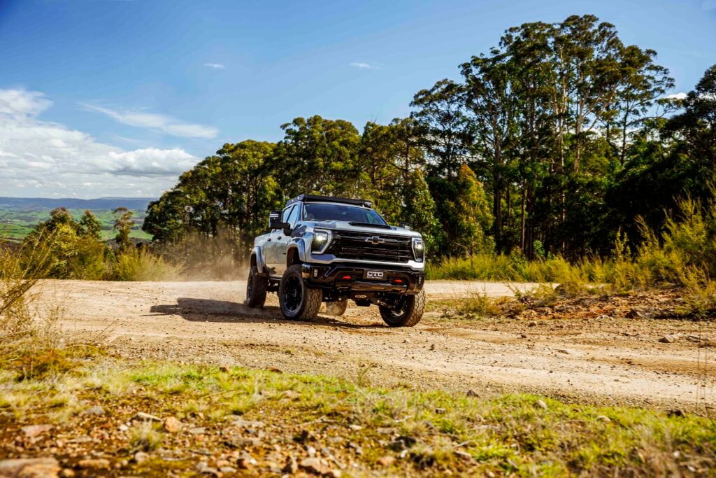White lifted pickup truck kicking up dust on a rugged dirt trail with greenery and blue sky in the background