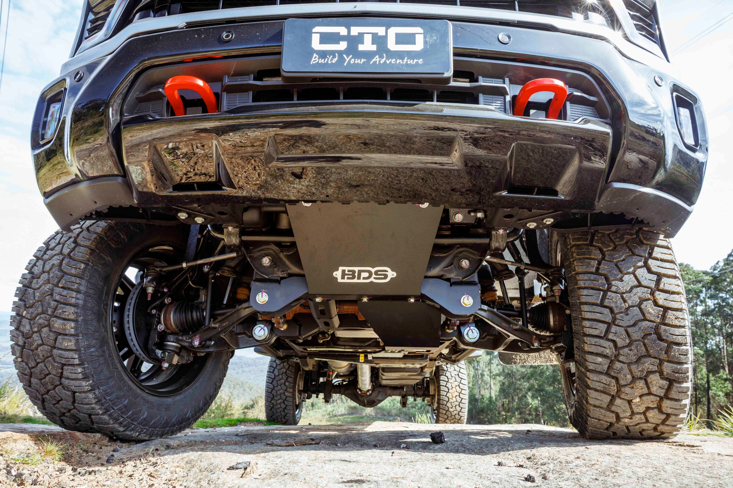 Low-angle view of a lifted off-road truck's underside, showing oversized mud-terrain tires, heavy suspension, and a BDS bumper/plating.
