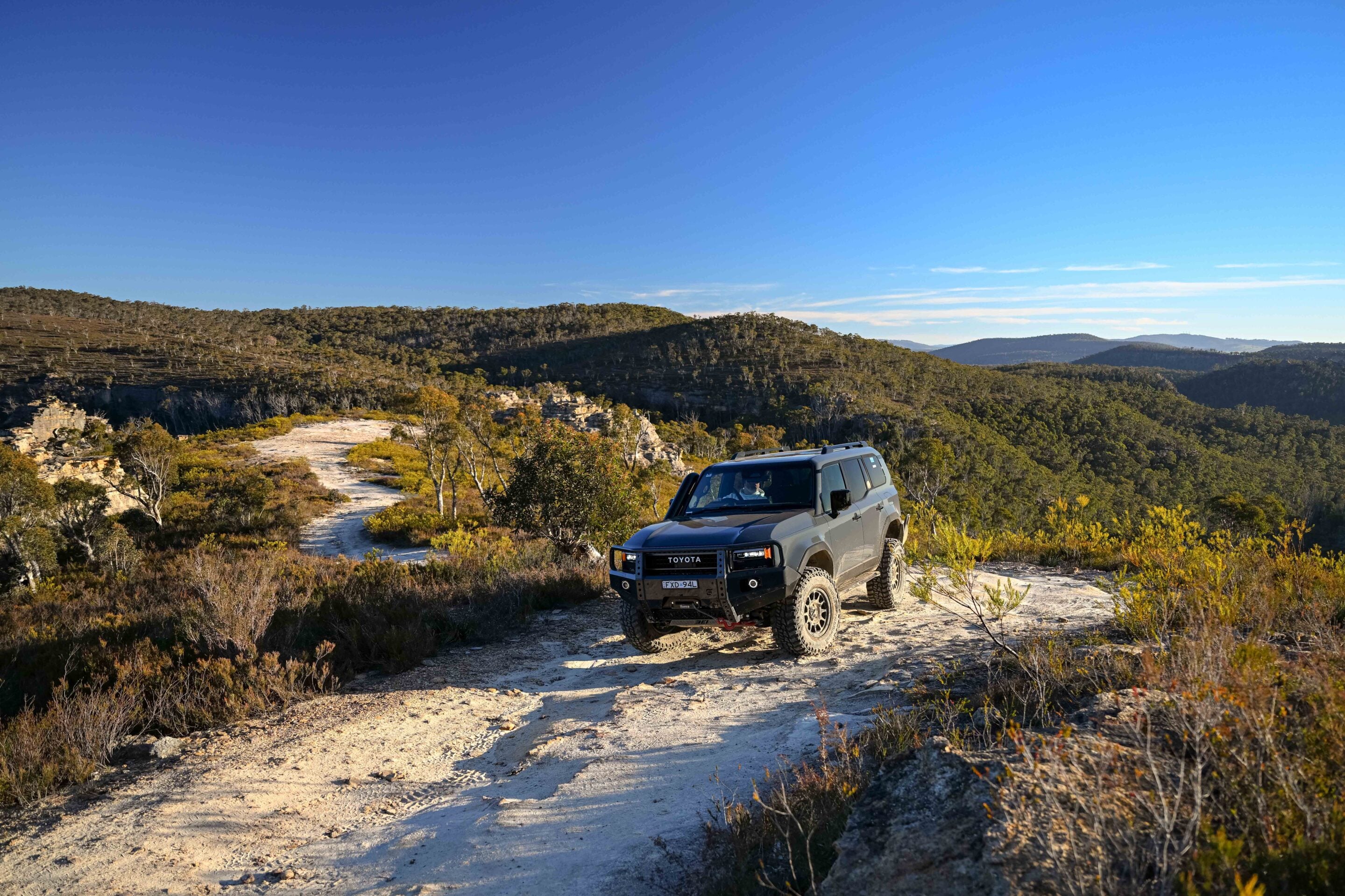 Gray Toyota 4x4 SUV on a rocky dirt trail in a dry, hilly landscape with scrubby vegetation and a clear blue sky
