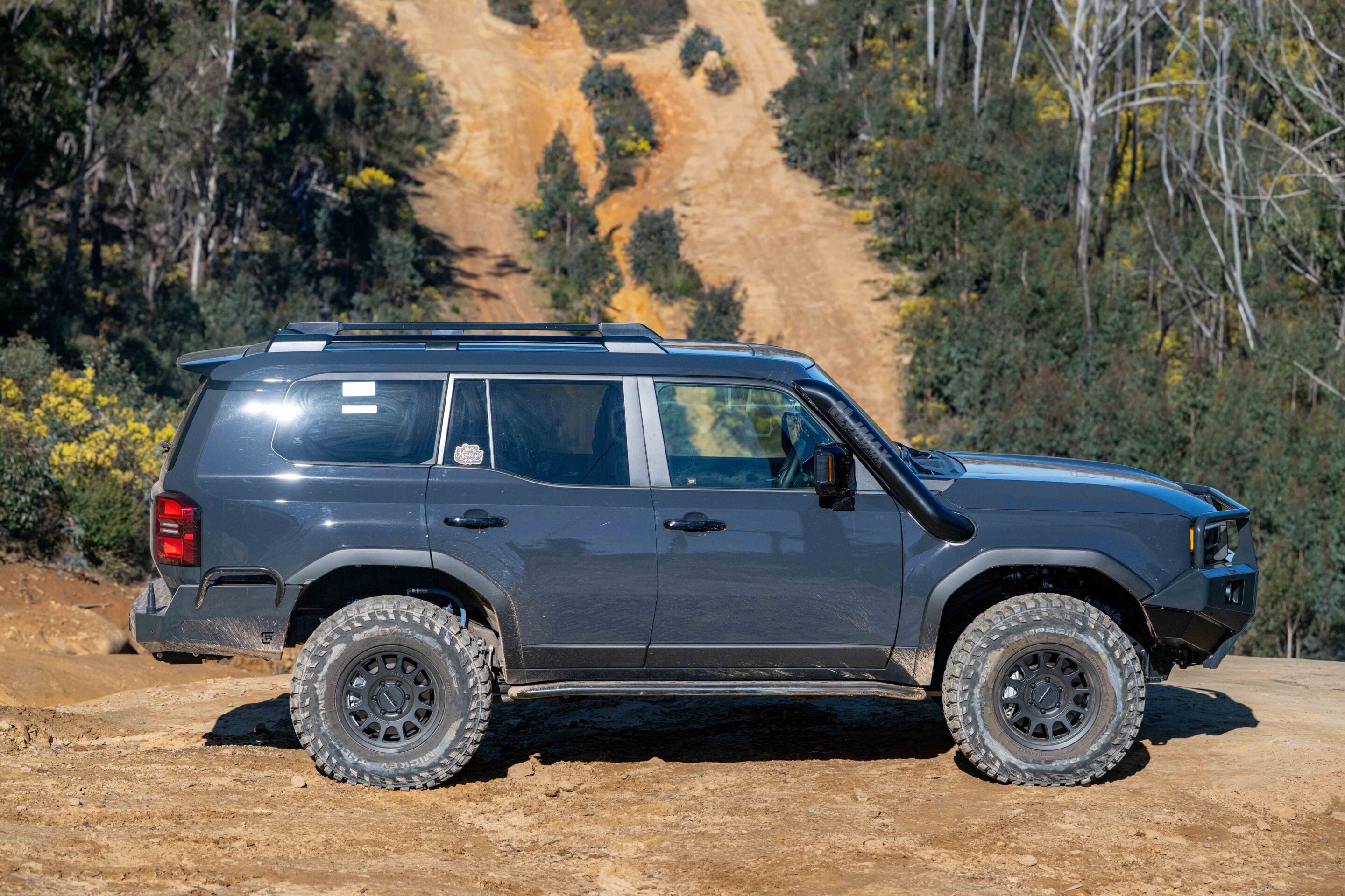 Side view of a gray off-road SUV parked on a dirt trail with a forested hillside in the background, rugged tires visible.