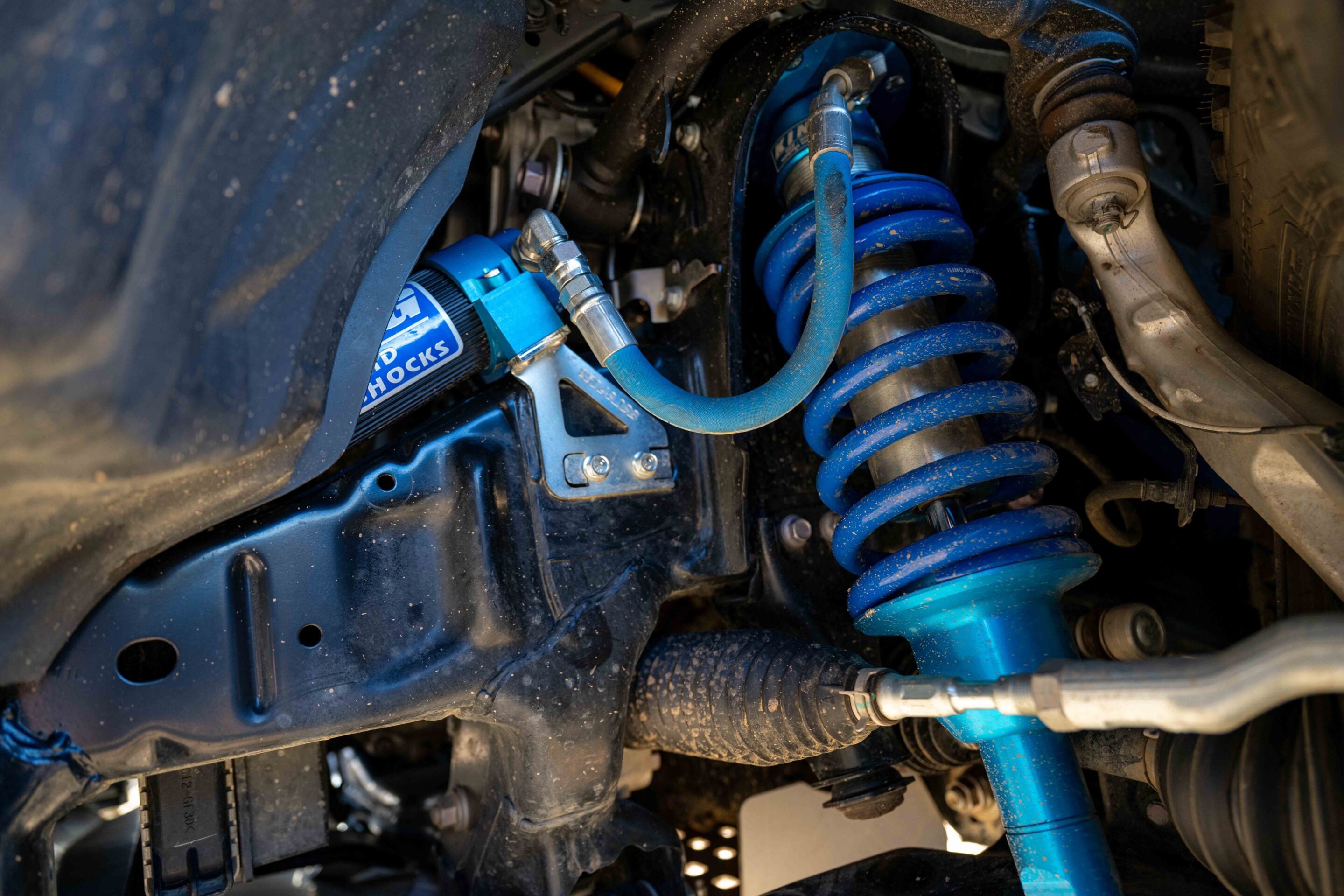 Blue coilover suspension with visible hoses and metallic fittings inside a dusty wheel well, part of a modified vehicle's rear suspension.