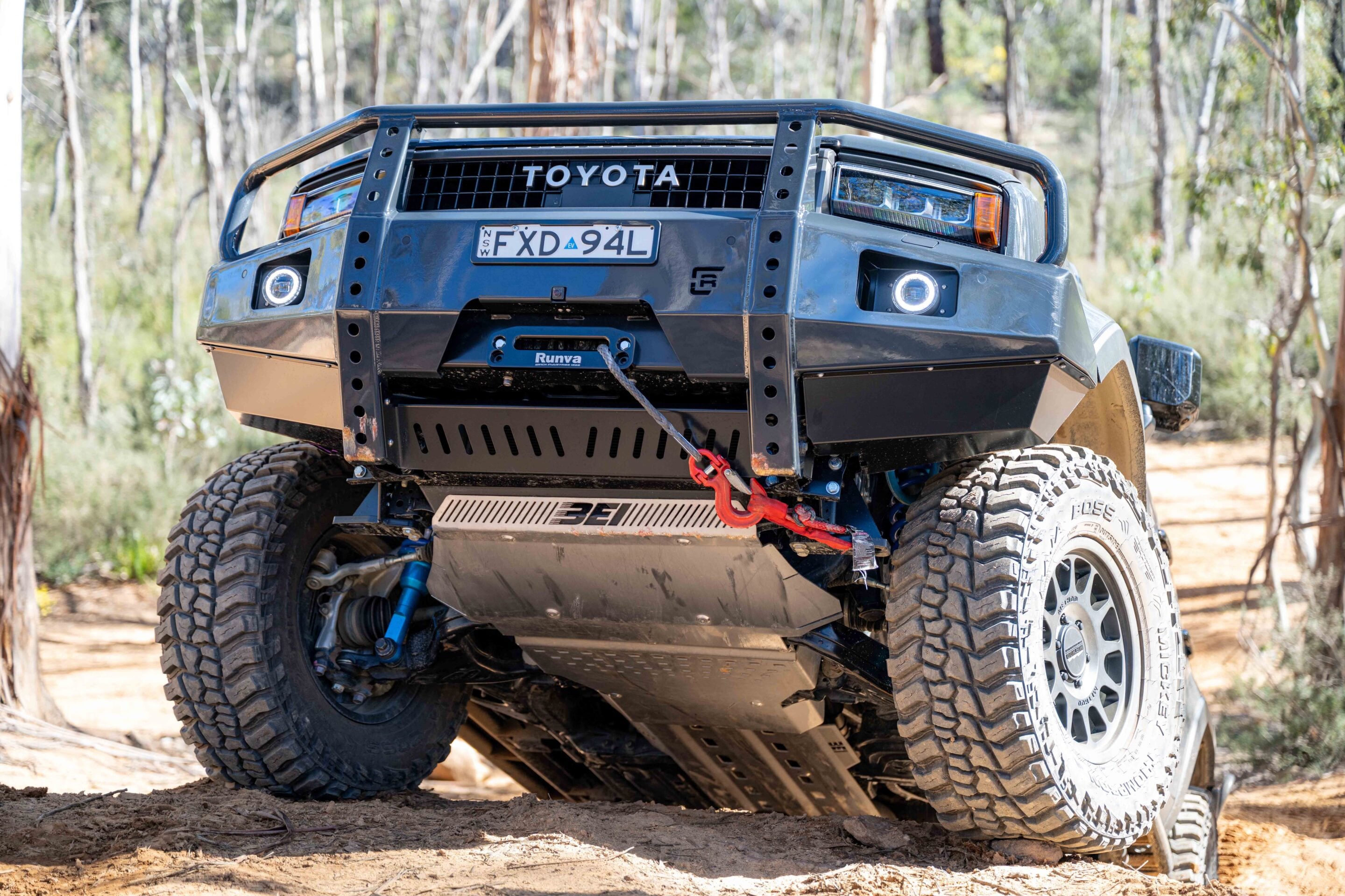 Front view of a modified off-road Toyota 4x4 with large mud tires and a red tow hook on a forest trail