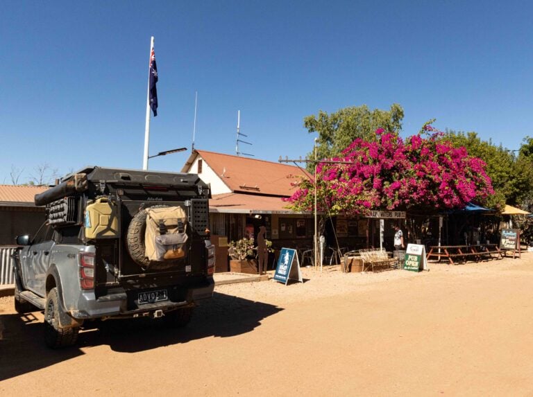 Rear of a rugged black SUV with gear mounted, parked on a dusty street beside a row of shops and pink bougainvillea.