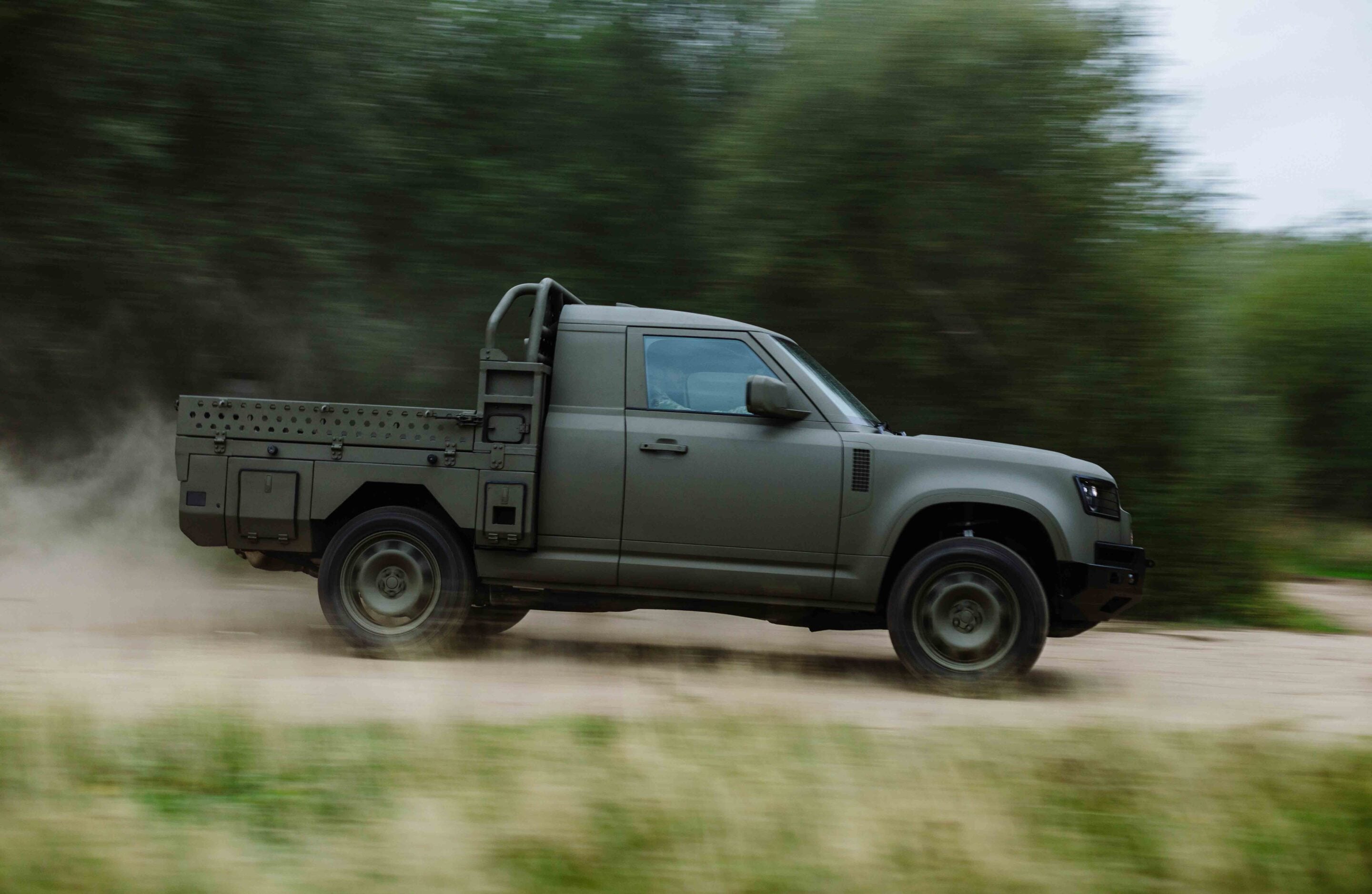 Green military-style pickup speeding along a dusty off-road trail with blurred trees in the background
