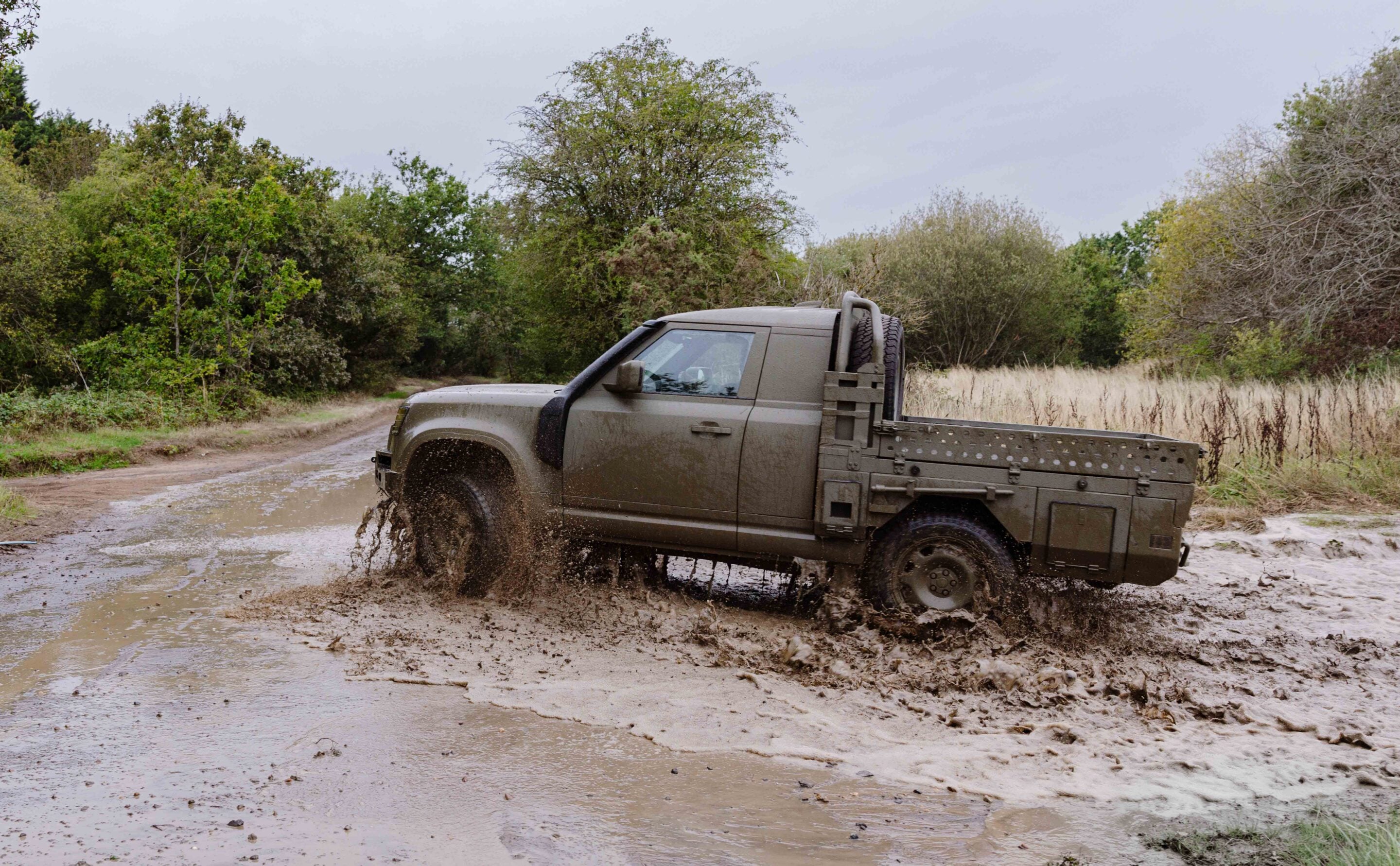 Off-road pickup truck driving through a muddy dirt trail surrounded by trees and bushes, mud splashing from the wheels.