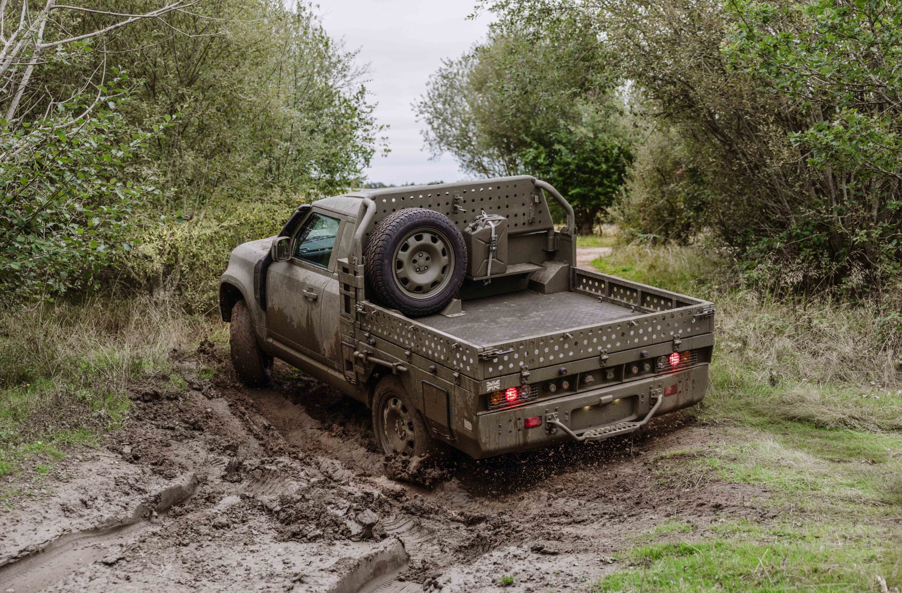 Mud-covered pickup truck with a spare tire in the bed, stuck on a muddy forest path surrounded by dense greenery.