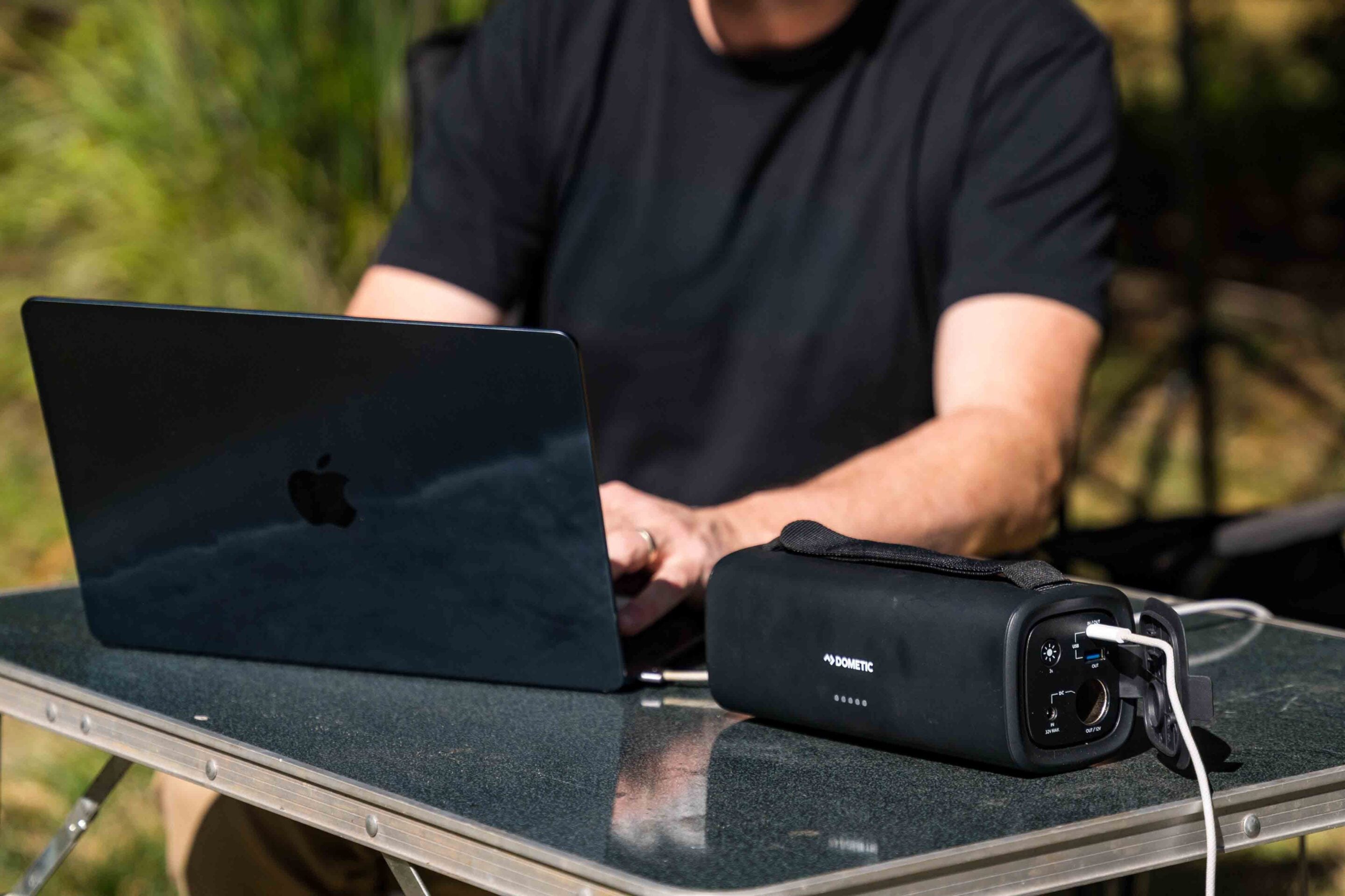 Person in a black T‑shirt typing on a laptop outdoors at a metal table, with a black portable power unit labeled Dometic nearby on the table.
