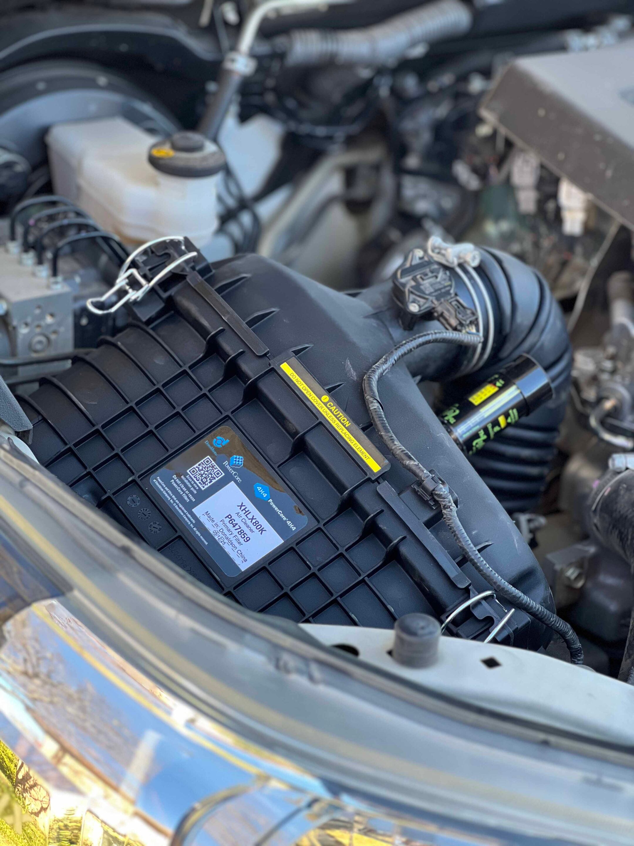 Close-up of a car engine bay showing a large black battery housing with wires and a white reservoir in the background.