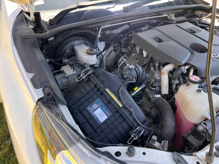 Front-left view of a car engine bay with the battery, coolant reservoir, and various hoses and wiring visible under the open hood