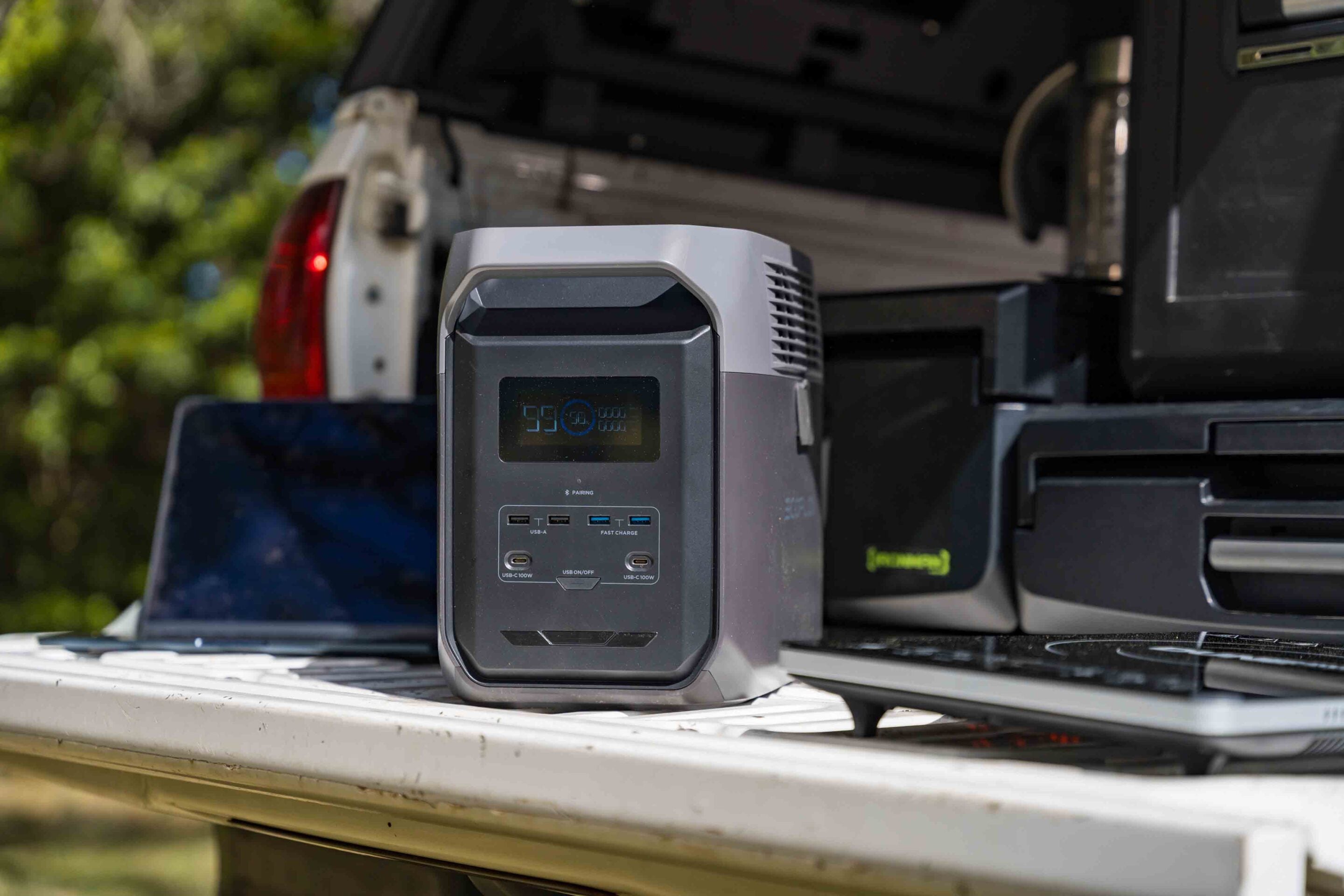 Portable power station with a digital display sits on the open tailgate of a pickup truck, showing USB ports and controls on its front panel.