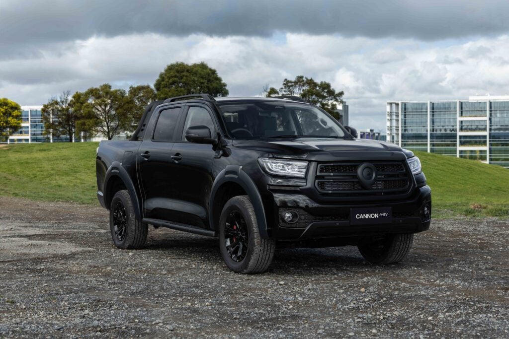 Black pickup truck parked on a gravel lot with modern office buildings and trees under a cloudy sky (plate reads CANNON PHEV).