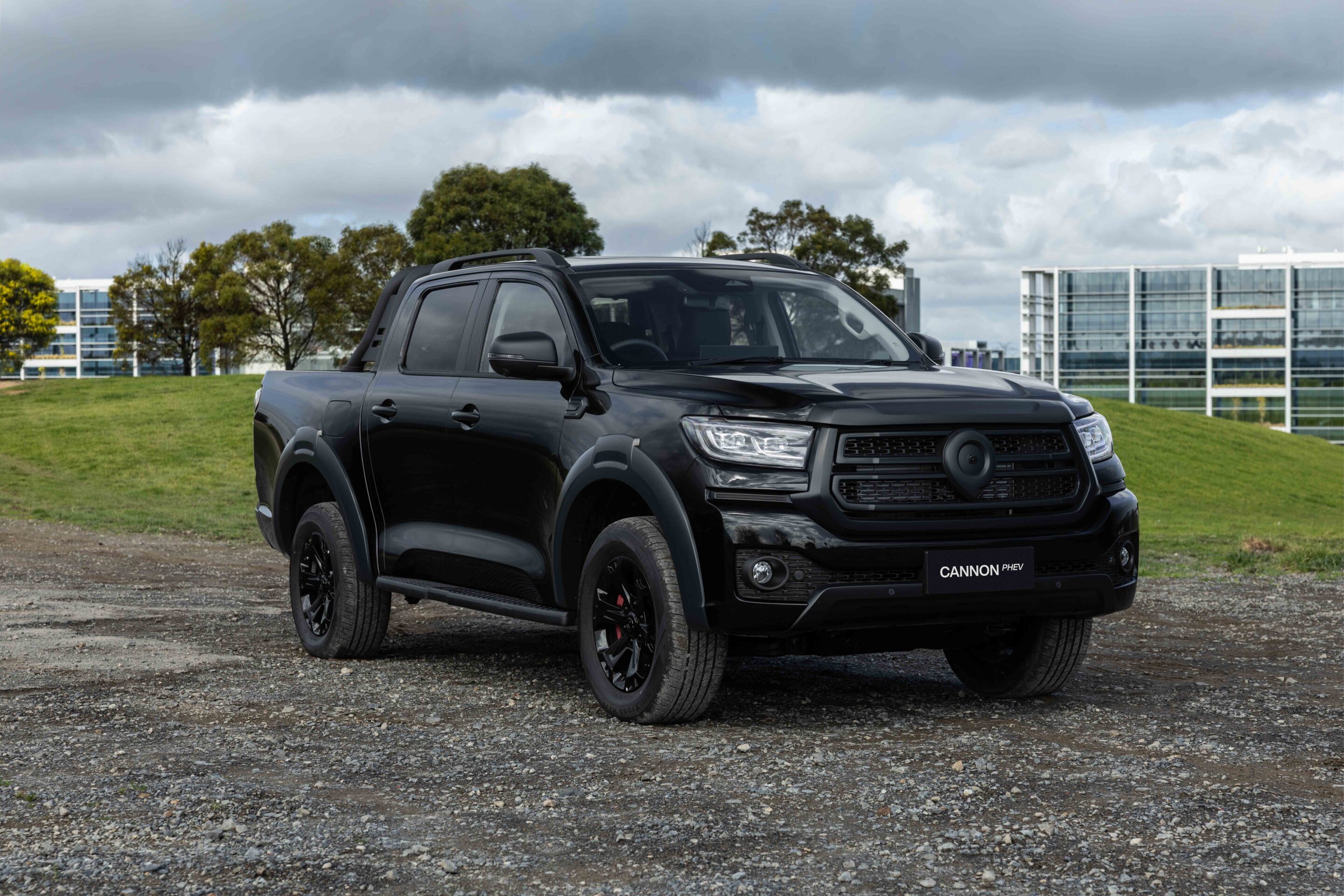 Black pickup truck parked on a gravel lot with modern office buildings and trees under a cloudy sky (plate reads CANNON PHEV).