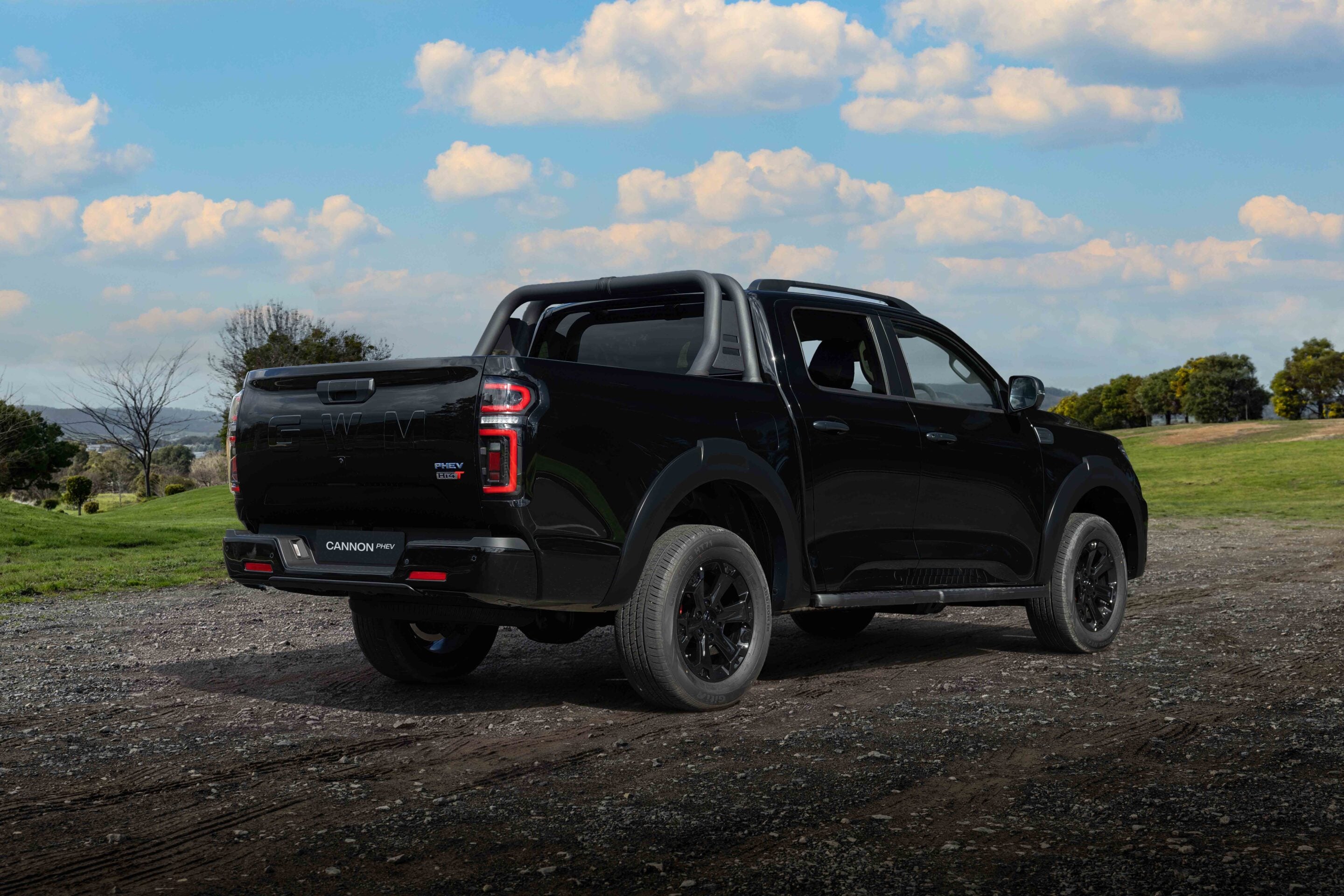 Rear-left view of a black pickup truck parked on a dirt/gravel road under a bright blue sky with fluffy white clouds, in a grassy landscapeóp