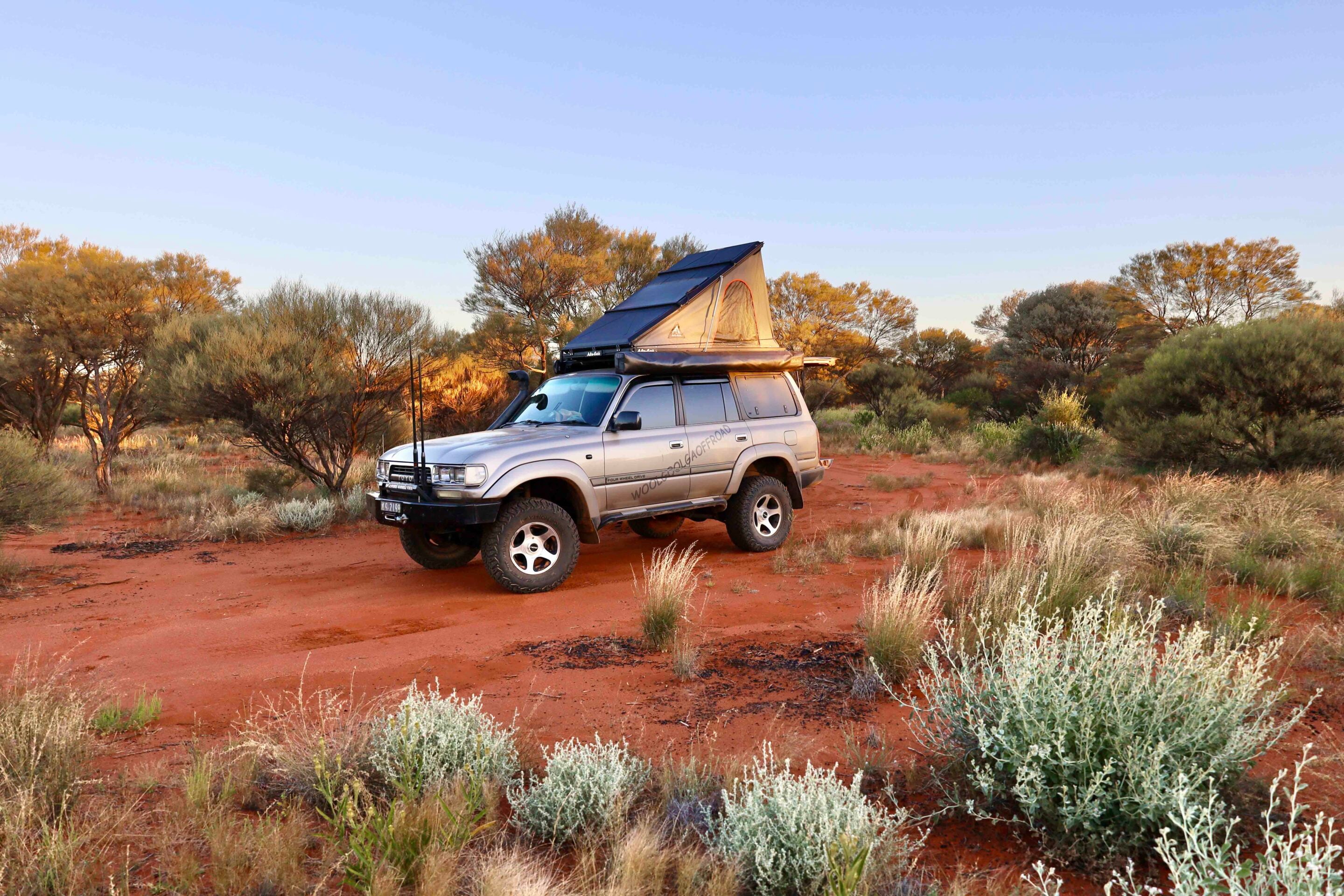 Silver SUV with a rooftop tent parked on red desert soil among scrubby bushes at sunset/sunrise caps off a camping setup