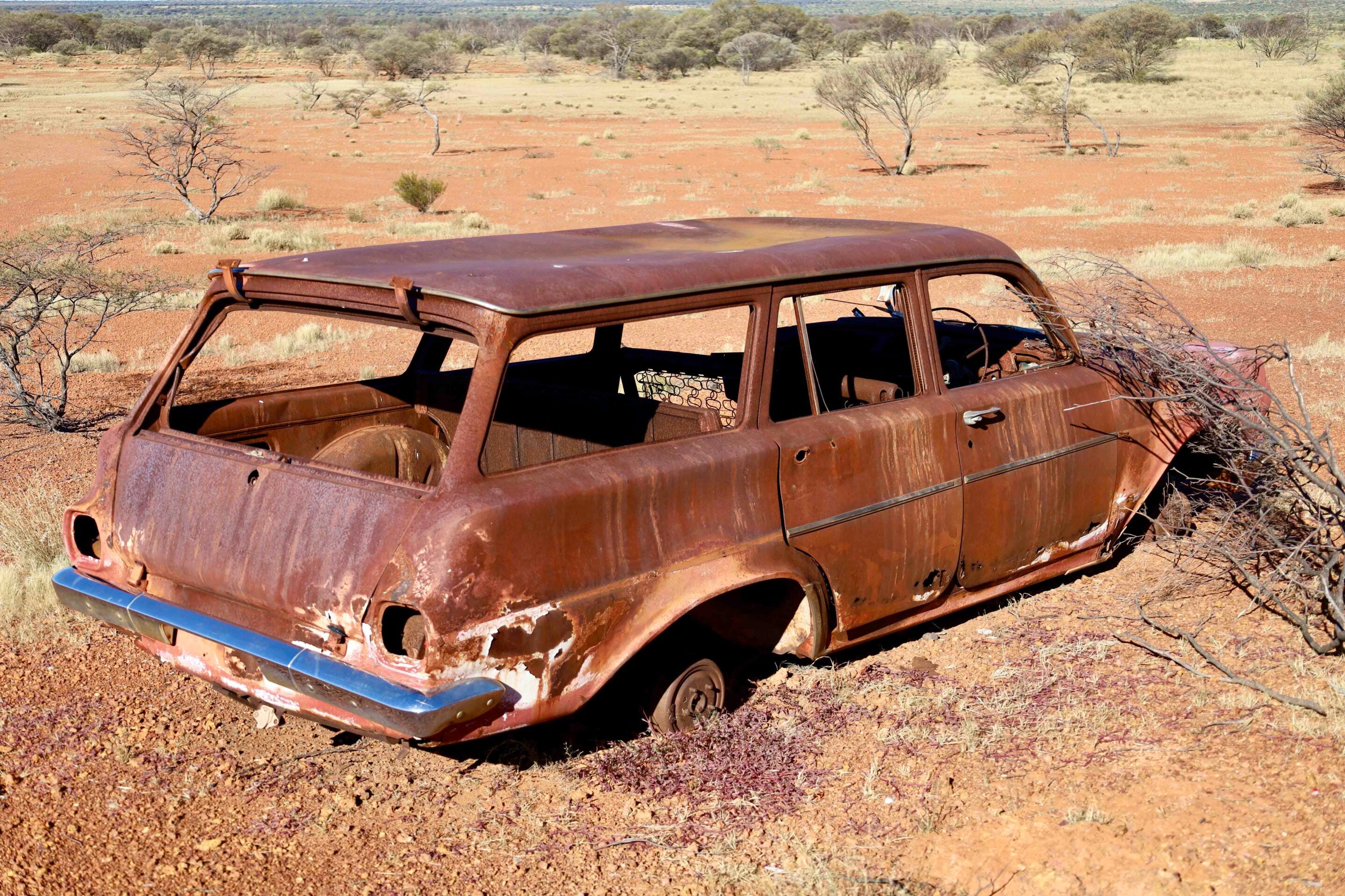 Rusty, abandoned car in a dry, red desert landscape with scattered shrubs and leafless trees in the background.
