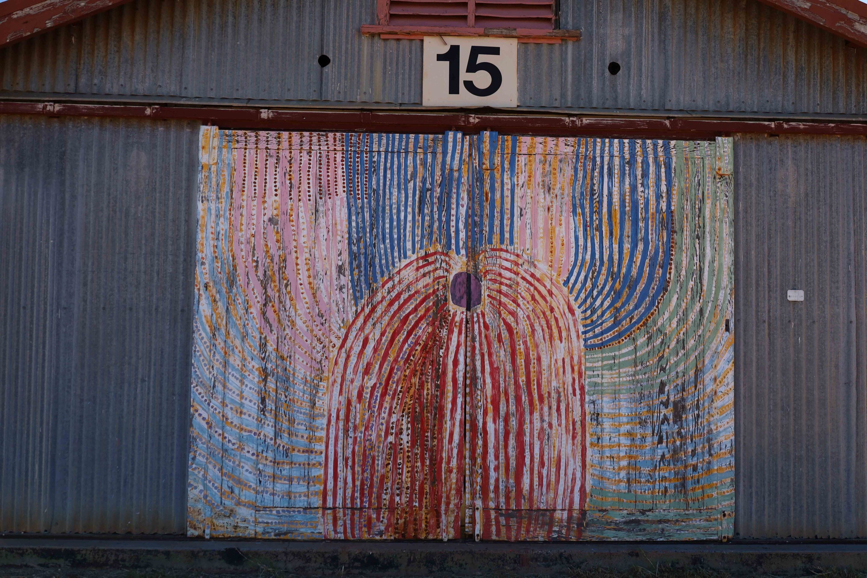 Rusty corrugated metal wall with a large colorful circular mural featuring rainbow stripes and a purple center; a white 15 sign is above the mural.