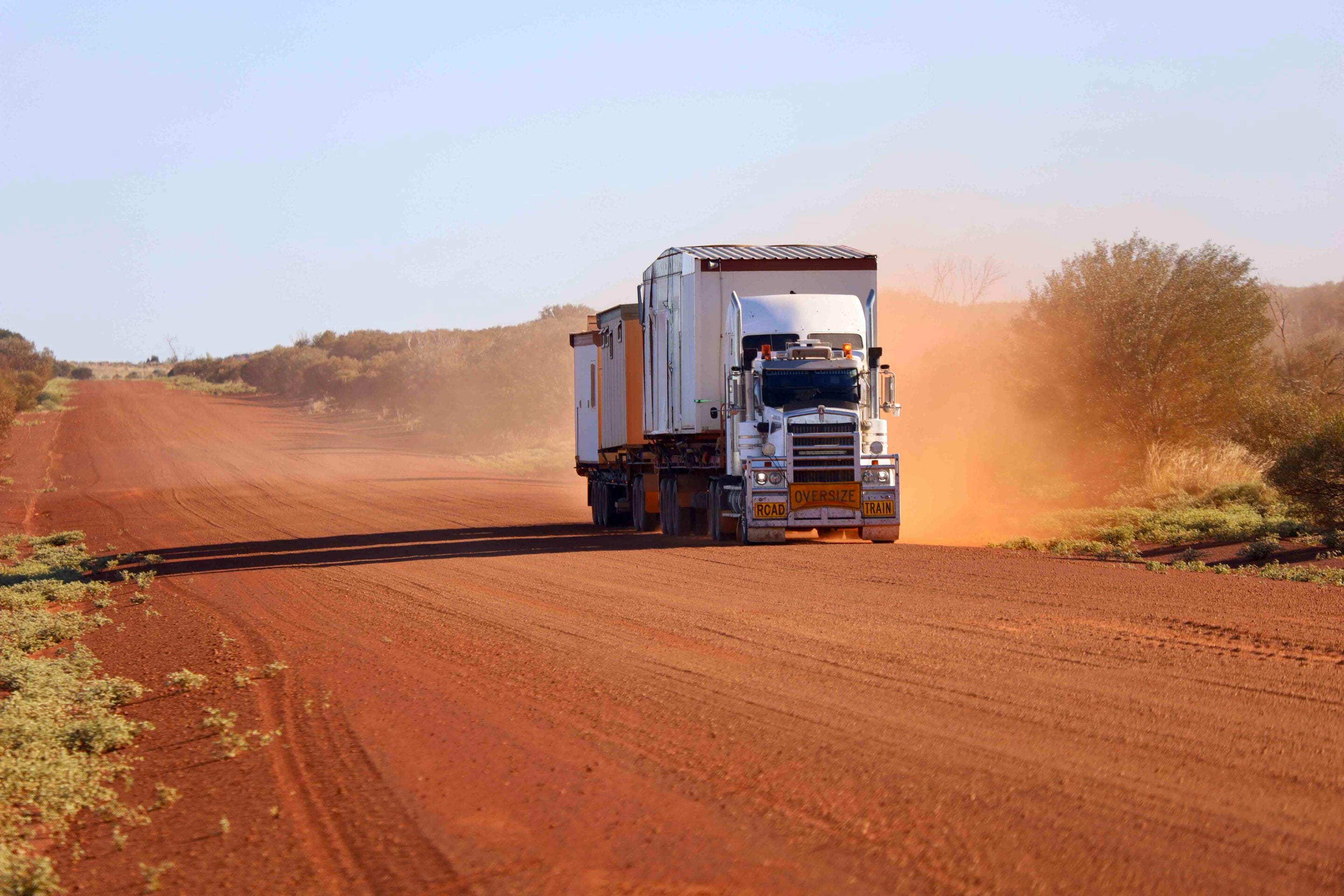 Oversize white semi truck hauling a long trailer on a dusty red dirt road in a desert landscape.