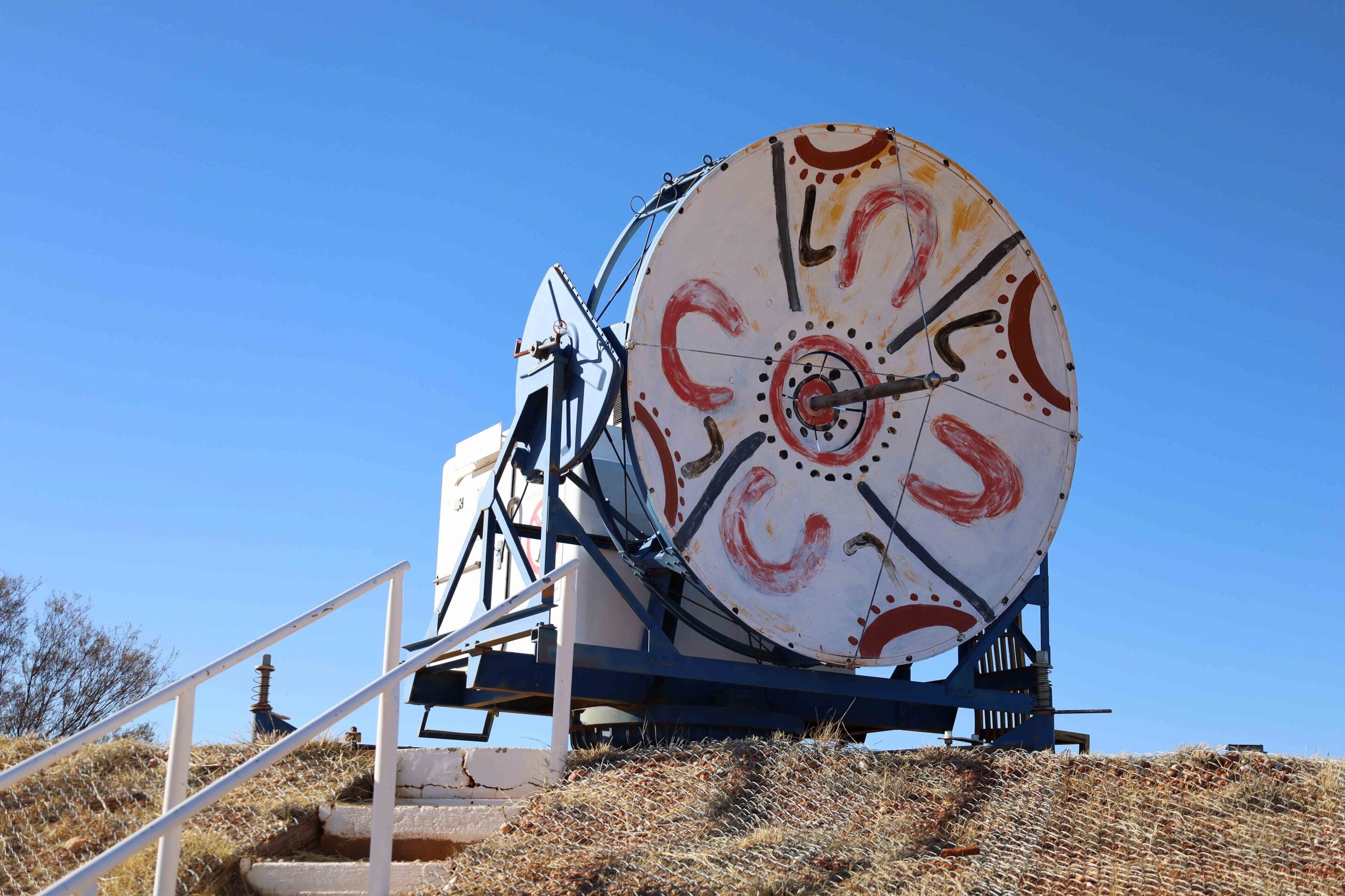 Large circular painted sign mounted on a blue metal structure against a clear blue sky, with a white railing and dry grass in the foreground.