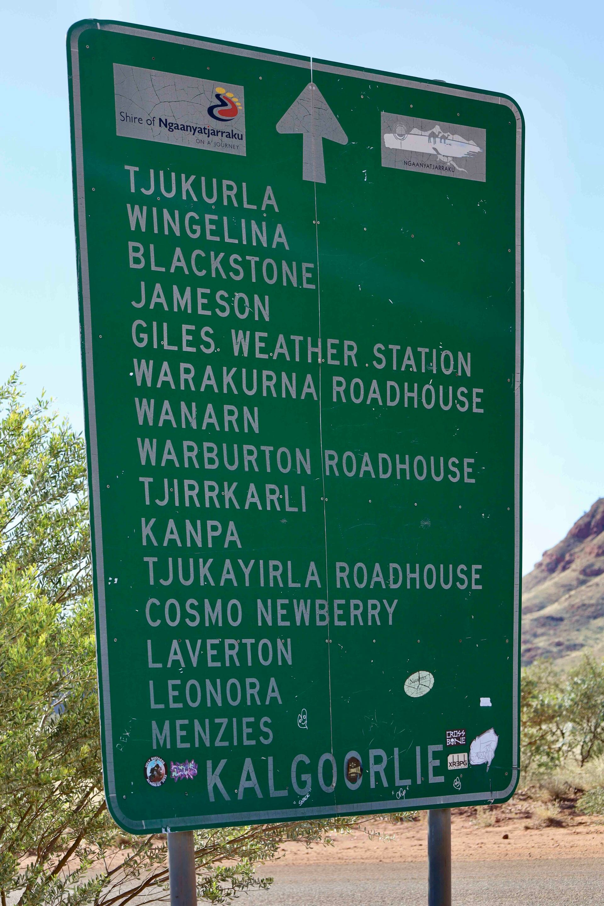 Green roadside sign listing many remote towns and roadhouses (e.g., Tjukurla, Wingelina, Blackstone, Jameson, Warburton Roadhouse, Kalgoorlie) with stickers, in a desert landscape.
