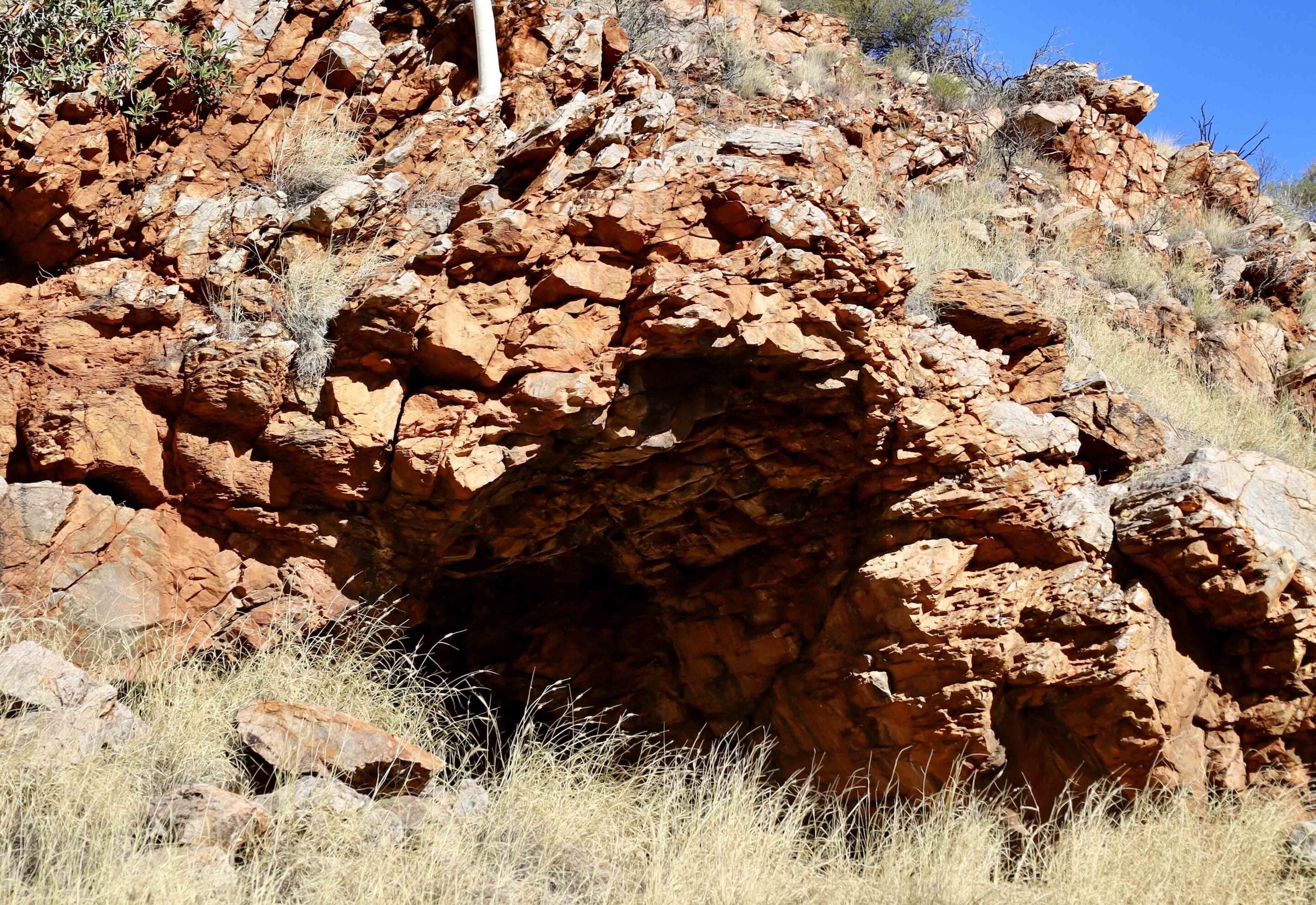 Rugged red rock cliff with sparse dry grasses at the base under a bright blue sky.