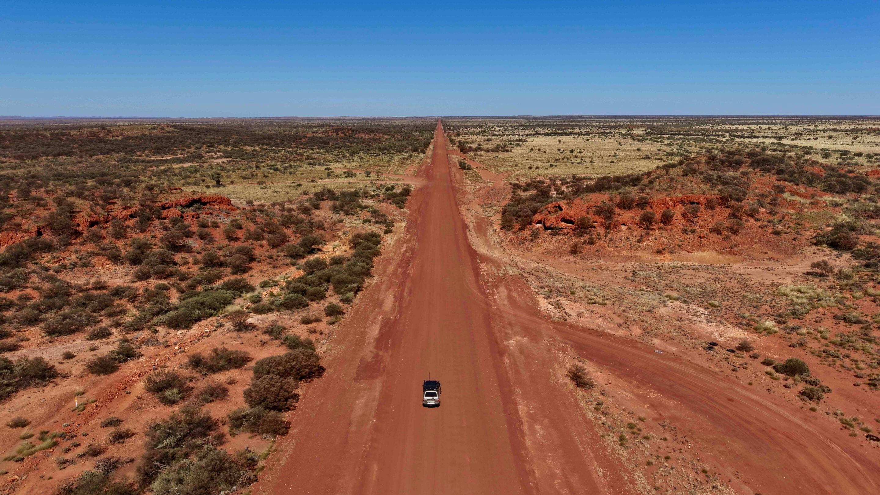 Aerial view of a straight red dirt road crossing a desert with sparse shrubs, two cars near the bottom, and a clear blue sky at the horizon.