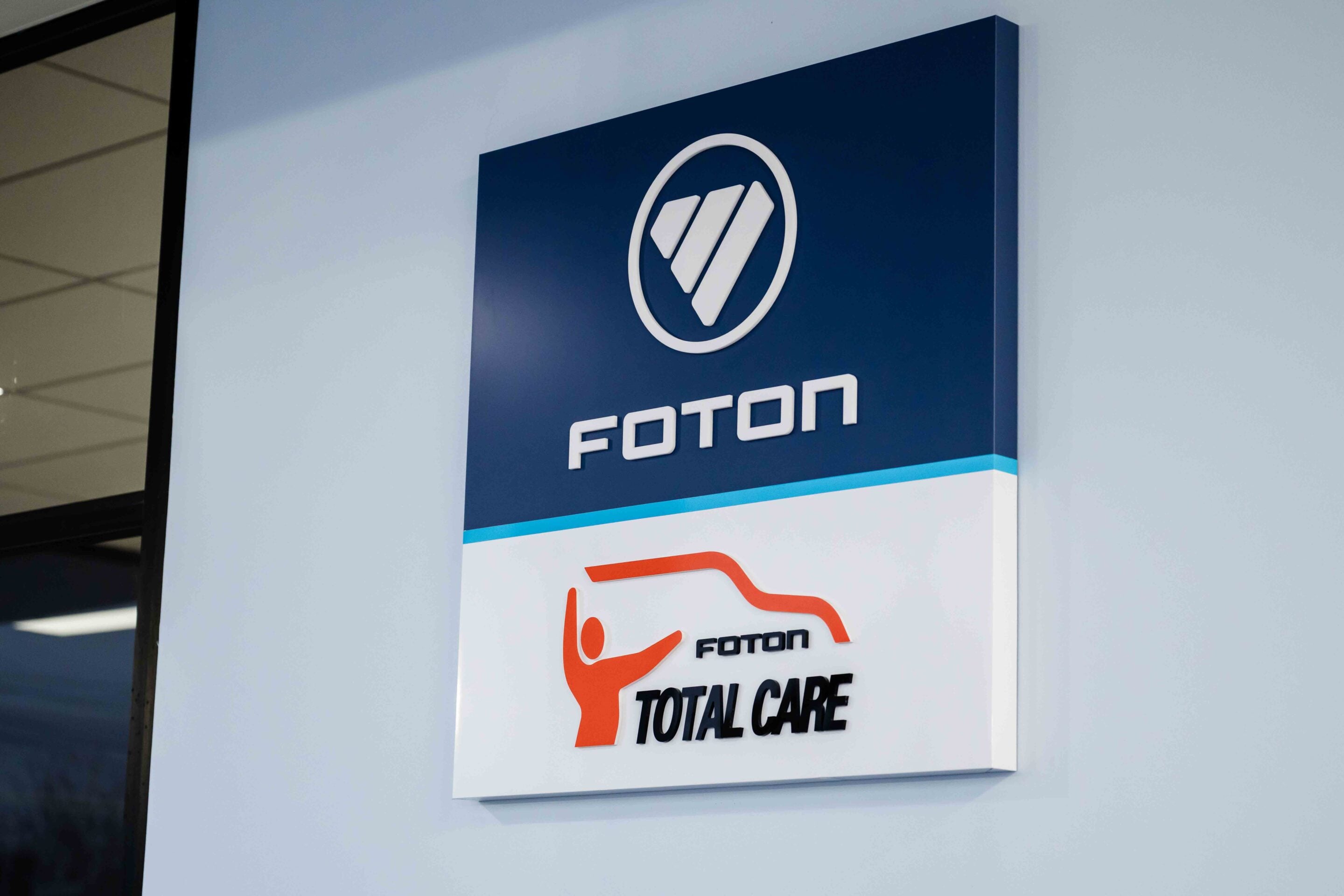 Blue wall sign featuring a white circular logo and the word FOTON, with an orange 'TOTAL CARE' graphic below.