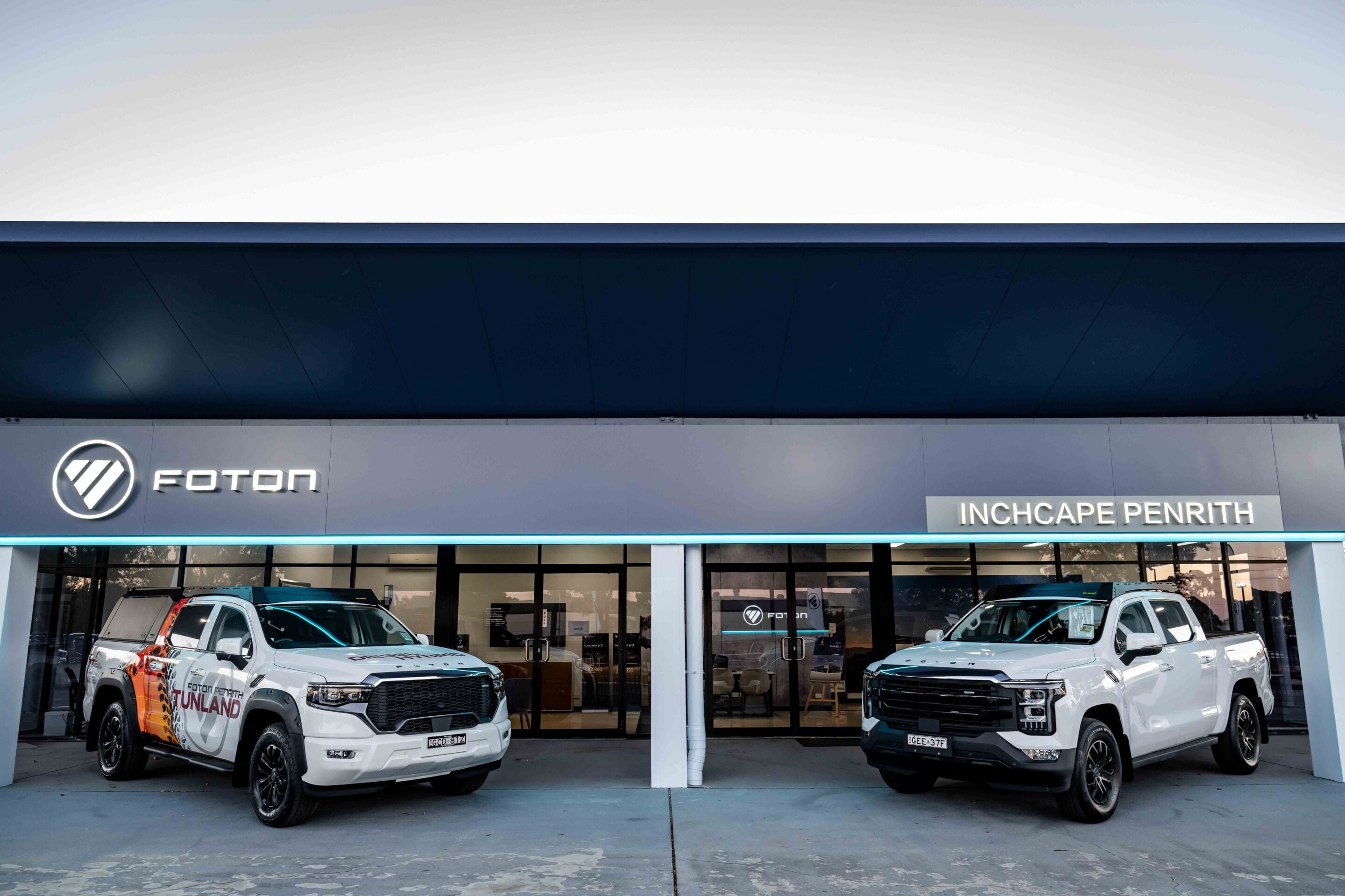 Front of a Foton dealership with two white pickup trucks parked outside under a dark overhang and a visible Foton logo.