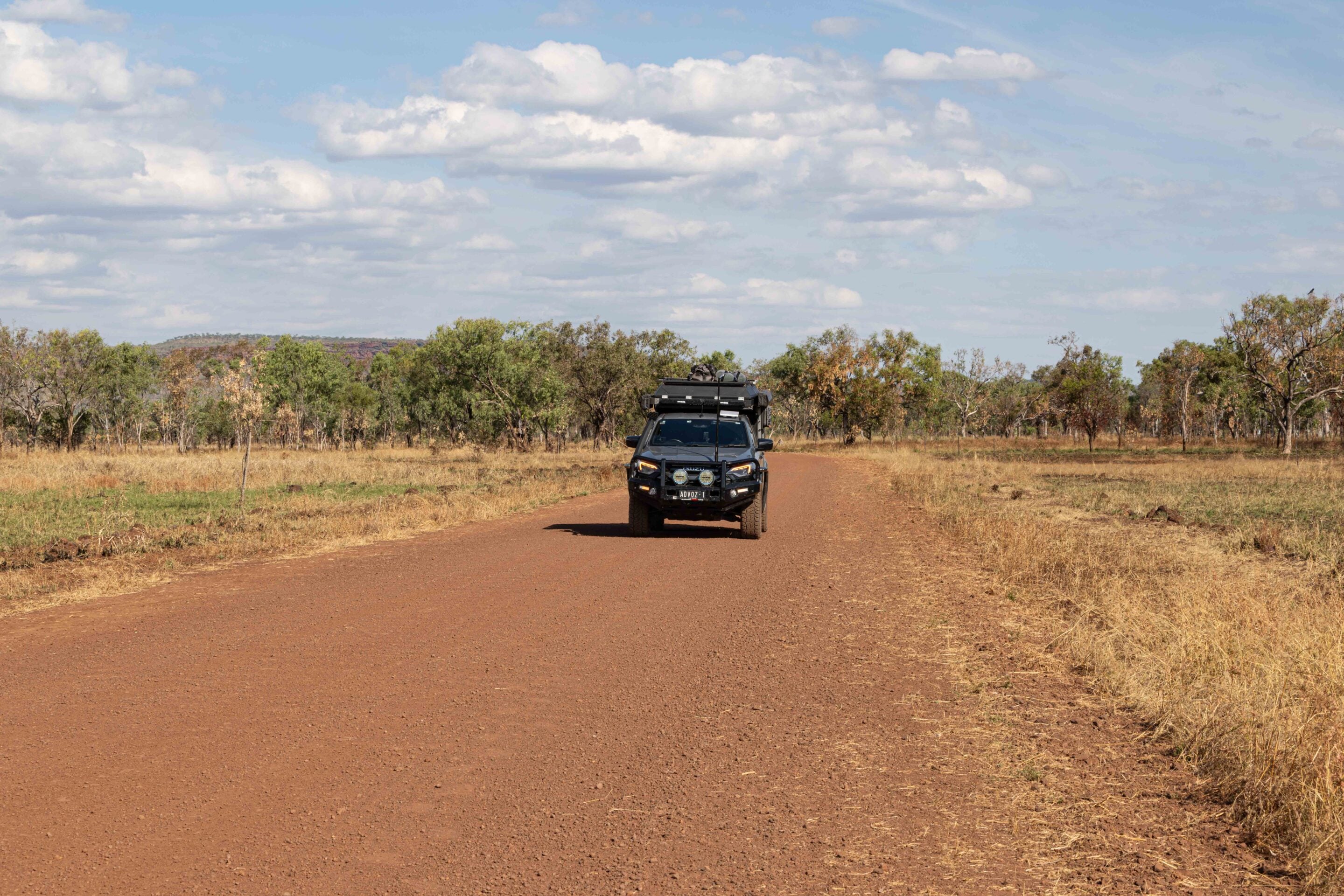 Off-road SUV with roof rack driving on a red dirt road through a dry savanna, blue sky and sparse trees ahead.