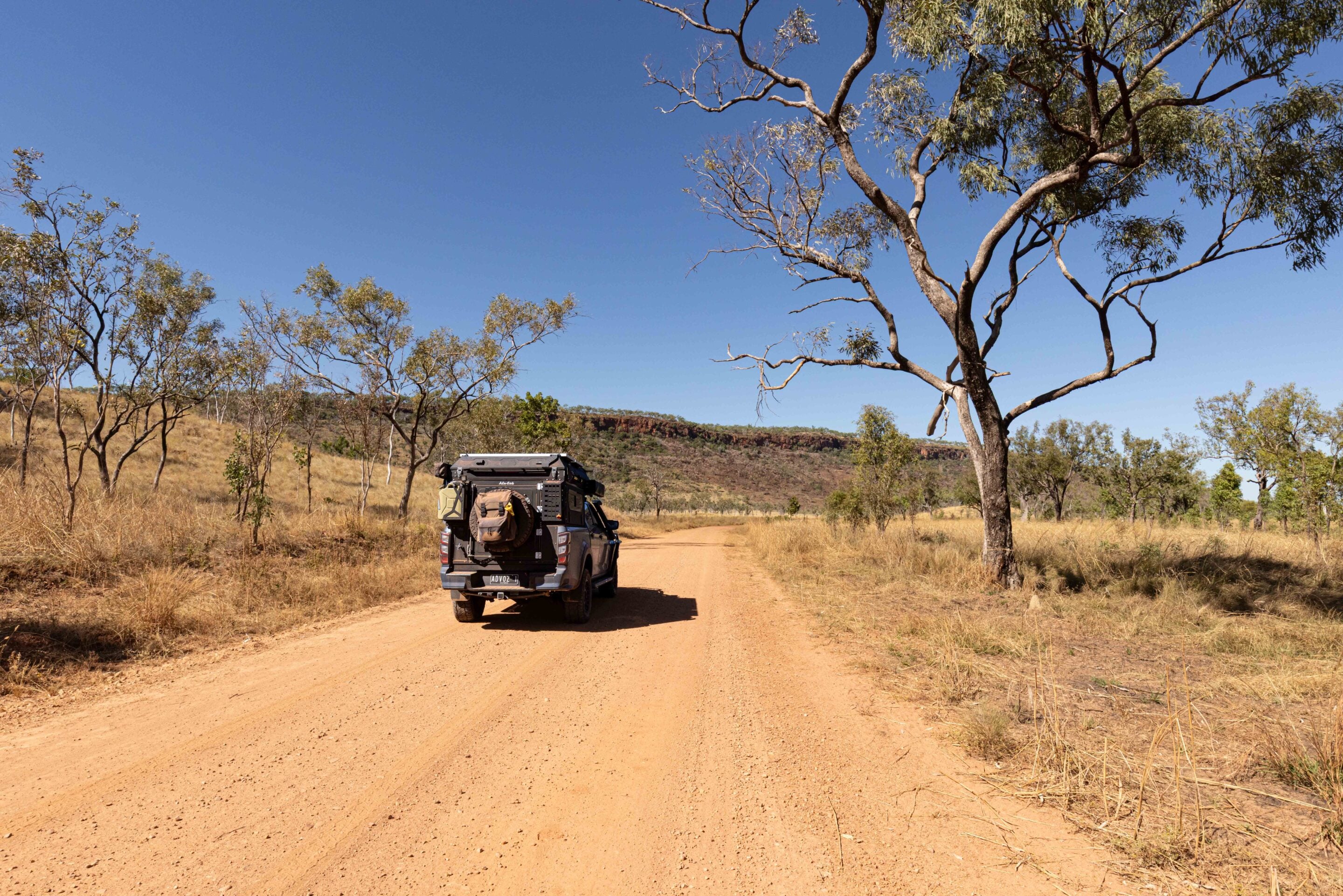 Off-road SUV with camping gear on a dusty dirt road in a dry landscape, with sparse trees and a clear blue sky.