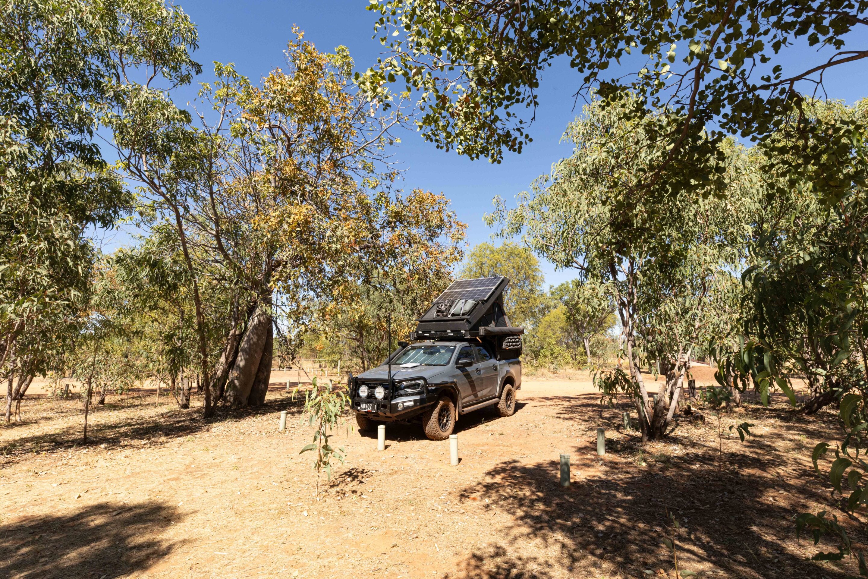 Grey off-road SUV with a roof-mounted solar panel and gear, parked among trees on a sunny day.