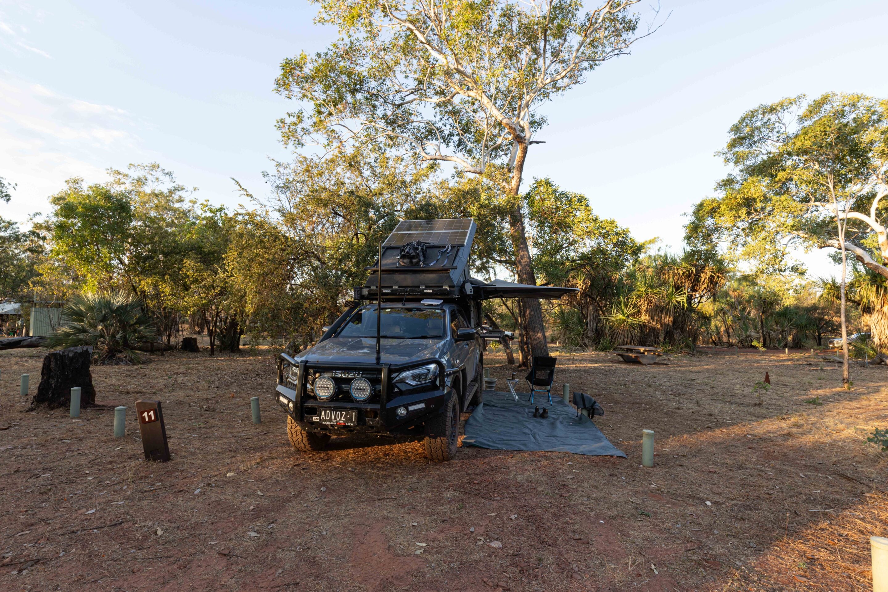 Isuzu SUV with a rooftop solar panel and camping gear at a dry woodland campsite.