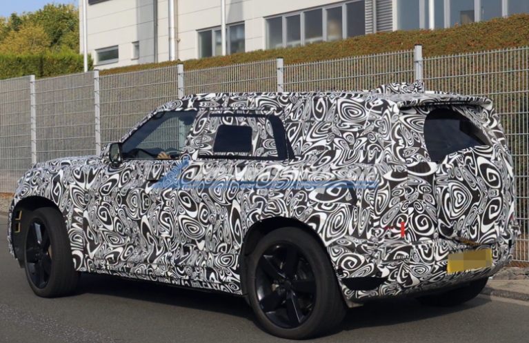 Camouflage-wrapped prototype SUV parked by a fence; black-and-white swirled wrap conceals design, rear plate blurred.
