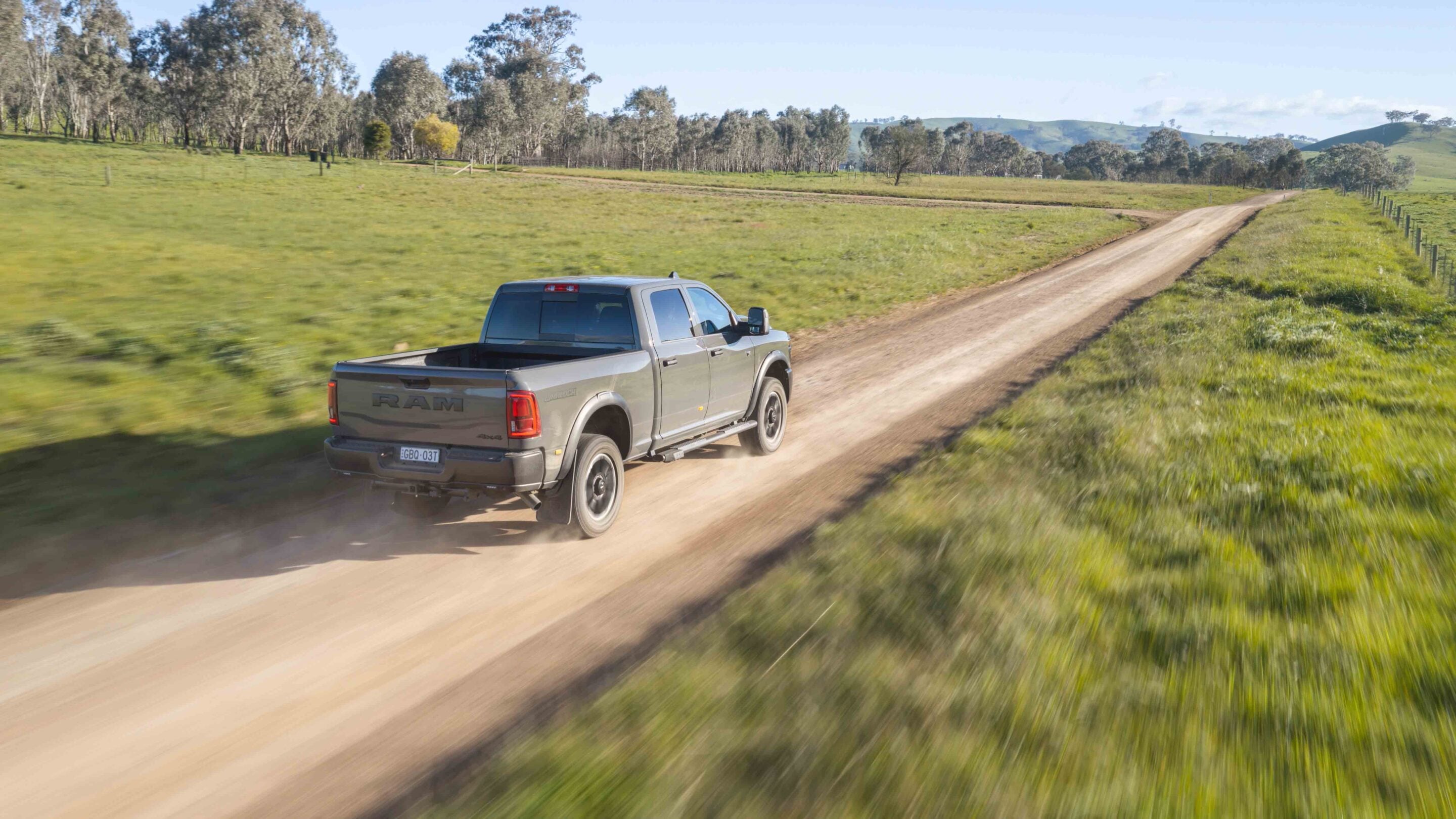 Silver RAM pickup driving on a dusty rural dirt road with green fields and trees to the left under a blue sky.