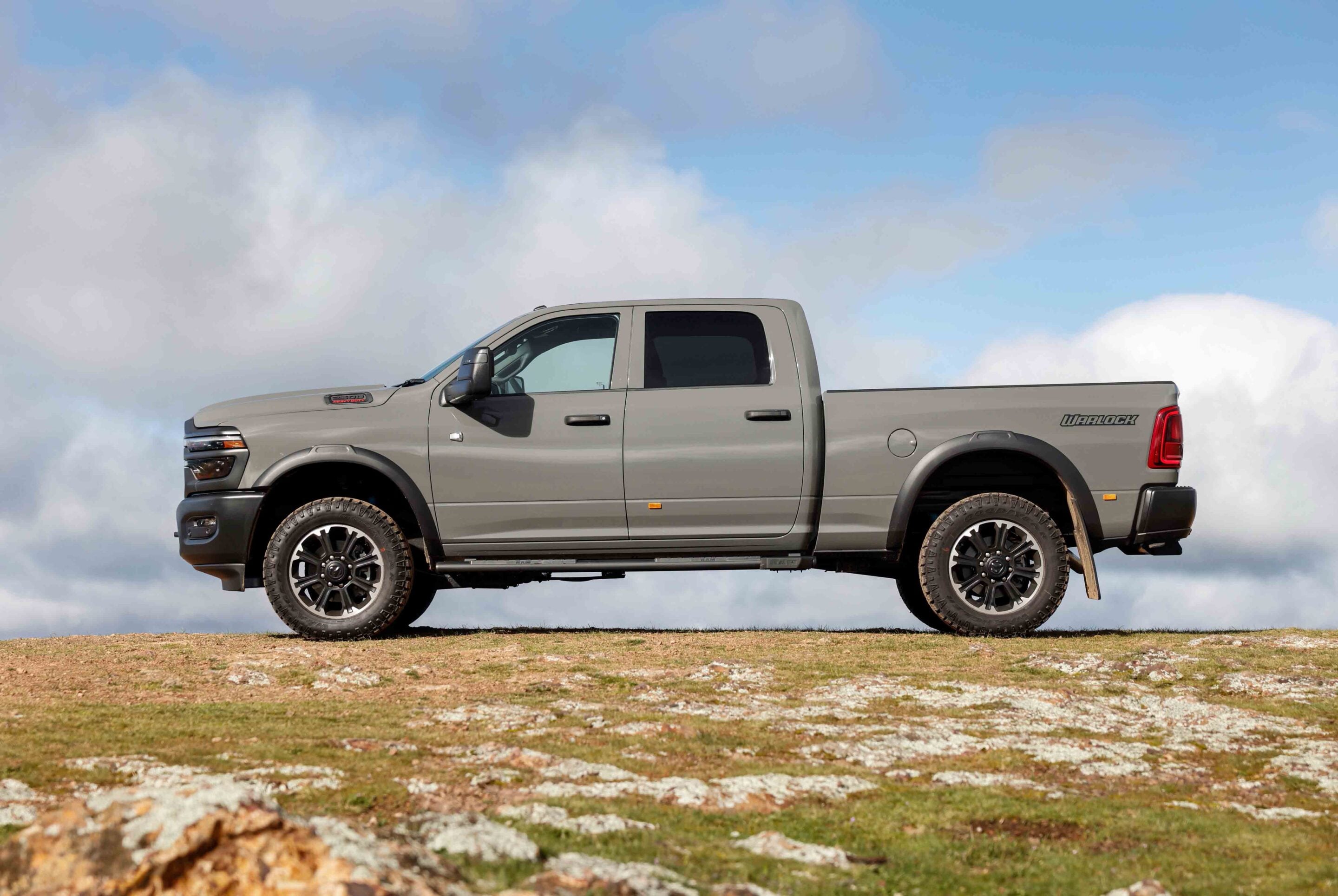 Side view of a gray pickup truck parked on a rocky dirt field under a cloudy blue sky.
