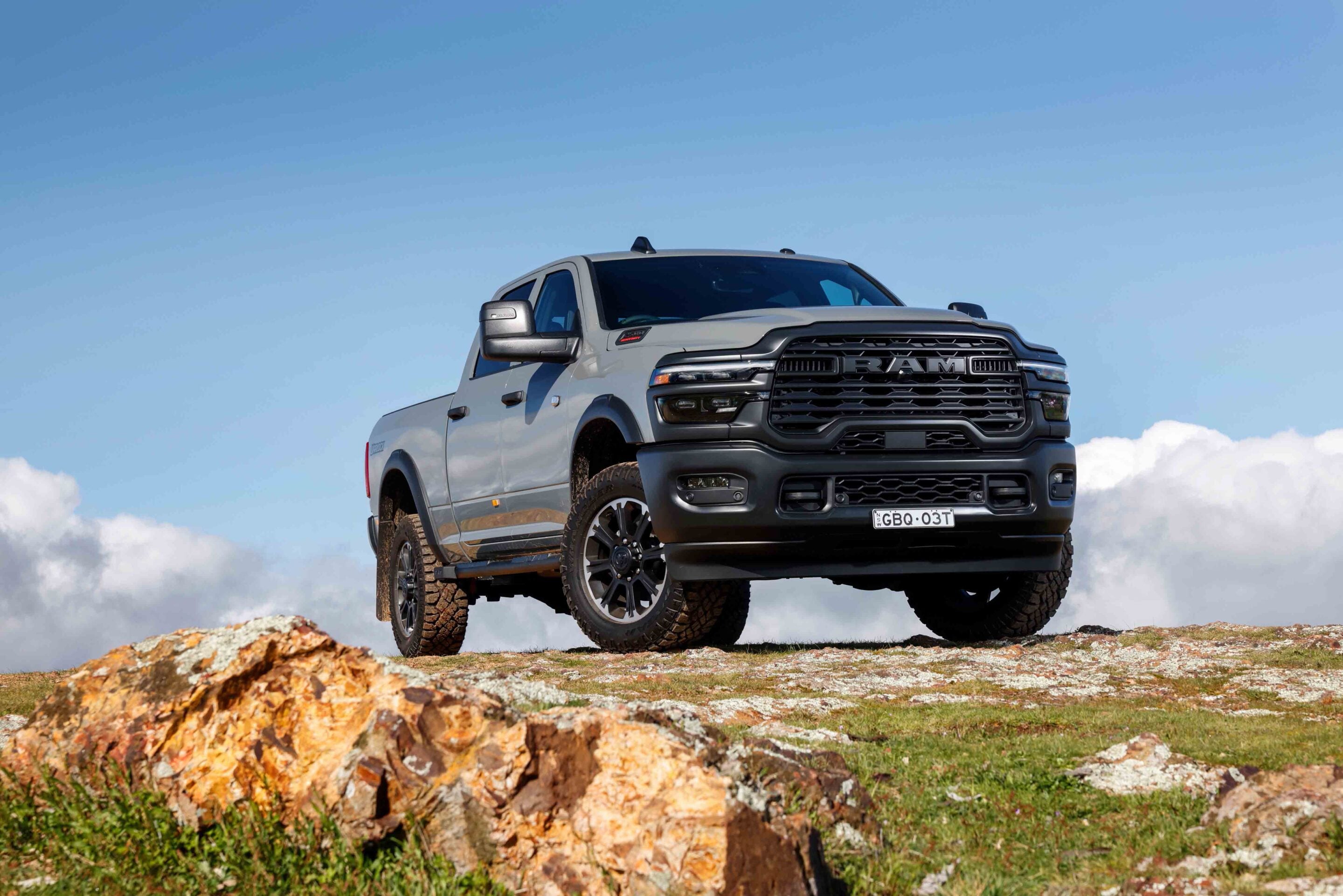 Silver RAM pickup truck perched on a rocky hill against a blue sky with clouds, front grille and large tires visible.