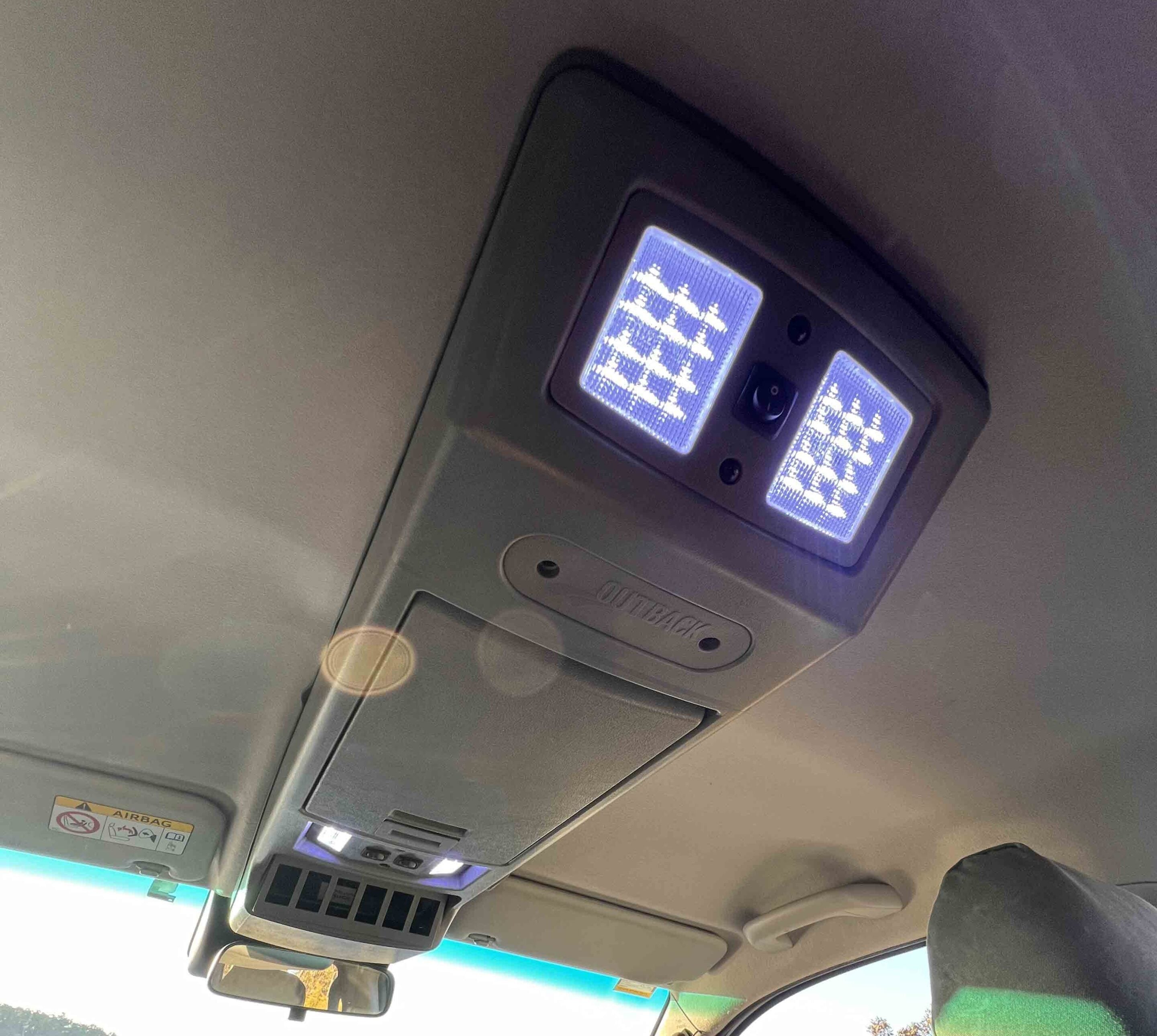 Overhead interior of a car with two illuminated ceiling lights and an overhead console, including an airbag warning label on the visor area.