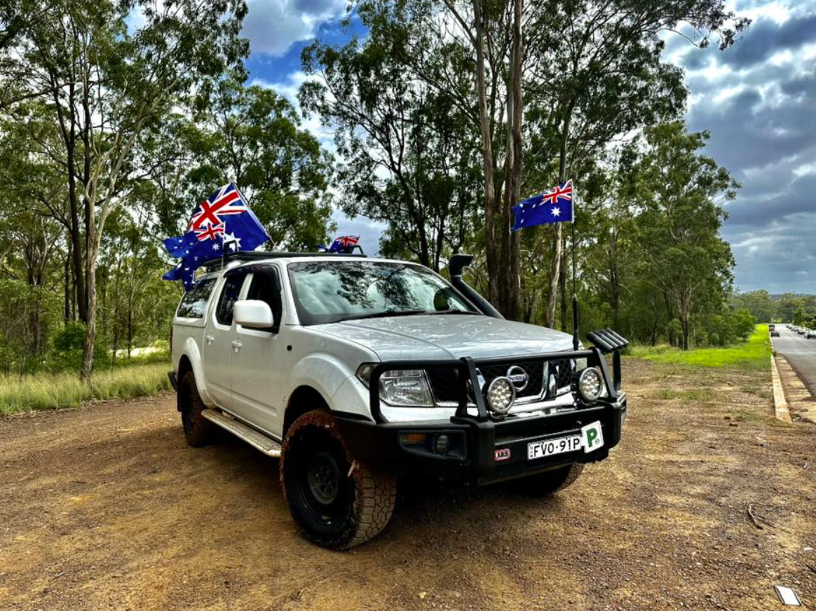 White off-road SUV with Aussie flags on the roof, parked on a dirt road among trees.