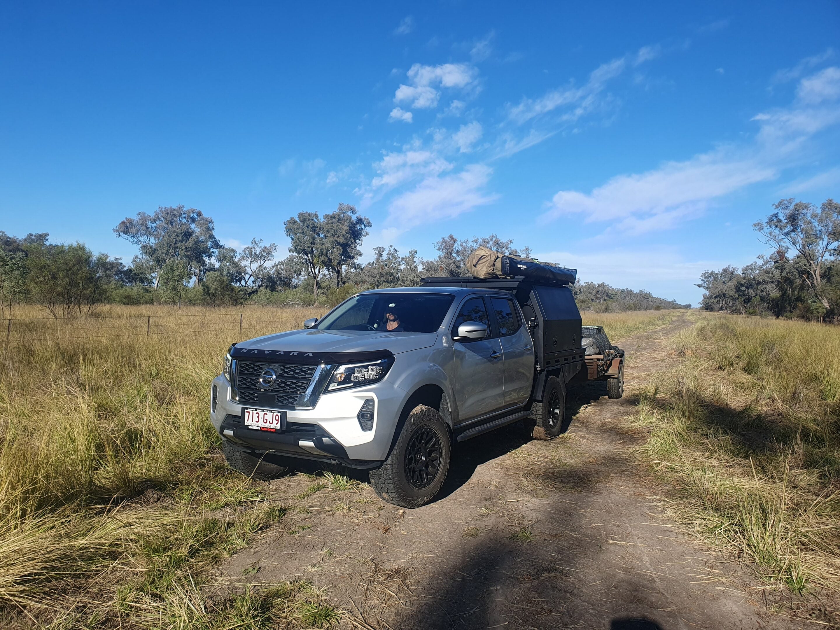 Silver off-road SUV with a roof-top camping tent parked on a dirt path in a grassy field under a bright blue sky.
