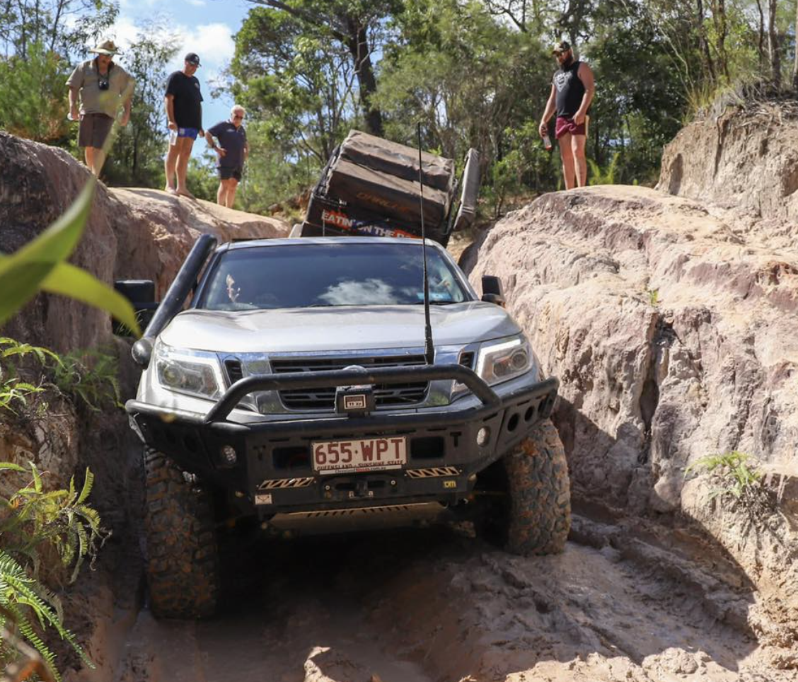 Silver off-road pickup wedged between rocky trench with large mud tires; several people stand on rocks watching the recovery.