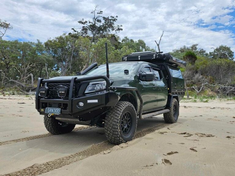 Dark green off-road pickup truck with a bull bar, snorkel, roof rack, and large mud tires parked on a sandy beach with trees in the background.