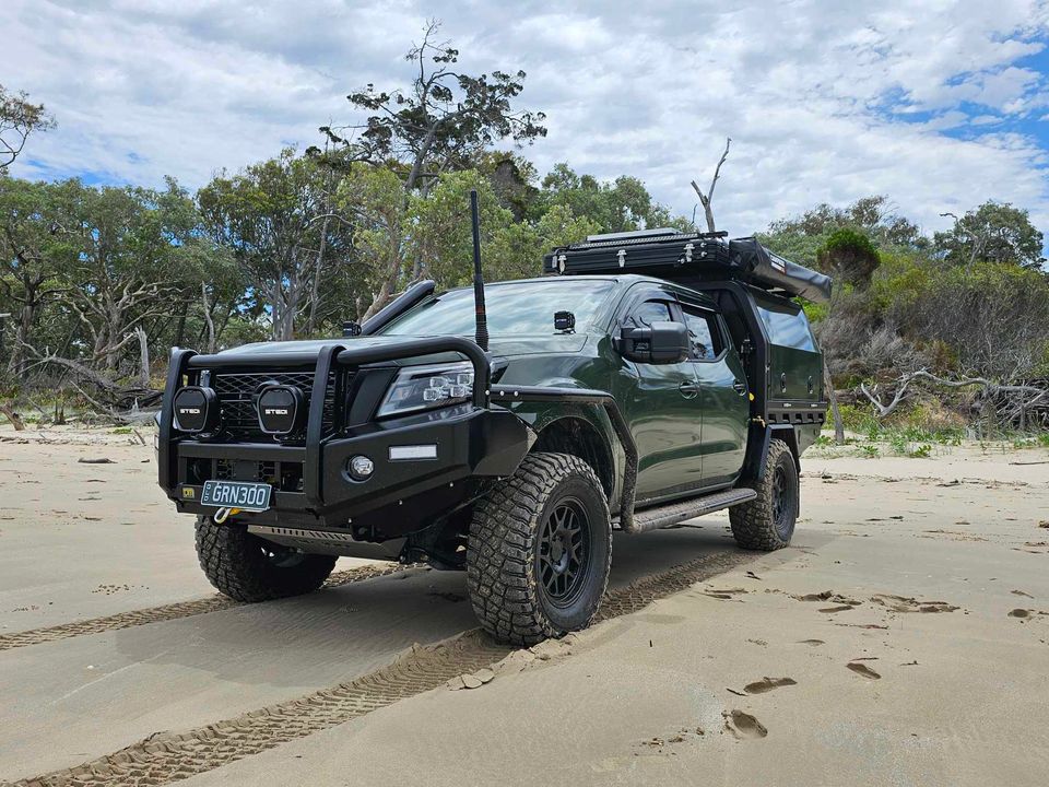 Dark green off-road pickup truck with a bull bar, snorkel, roof rack, and large mud tires parked on a sandy beach with trees in the background.
