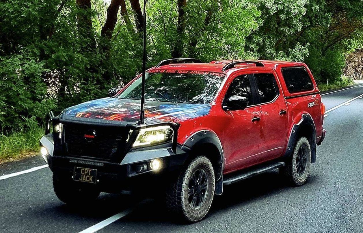 Red extended-cab pickup truck with a detailed hood graphic, off-road tires, and rust protection bars, driving on a forested road; license plate reads CPT JACK.