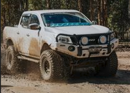 White pickup truck with large off-road tires driving through a muddy dirt trail in a forest.