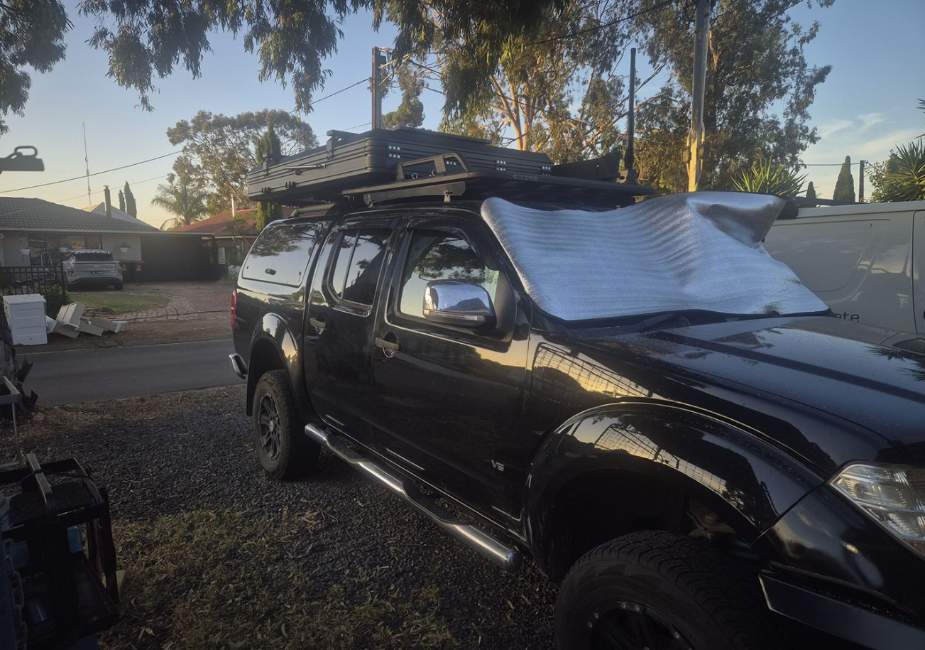 Black pickup truck parked in a driveway with a roof rack; a silver tarp covers the windshield area.