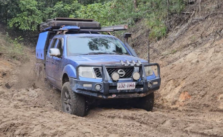 Blue off-road pickup truck driving through a muddy dirt trail with trees in the background, roof rack loaded with gear, front bumper guard visible (off-roading scene).