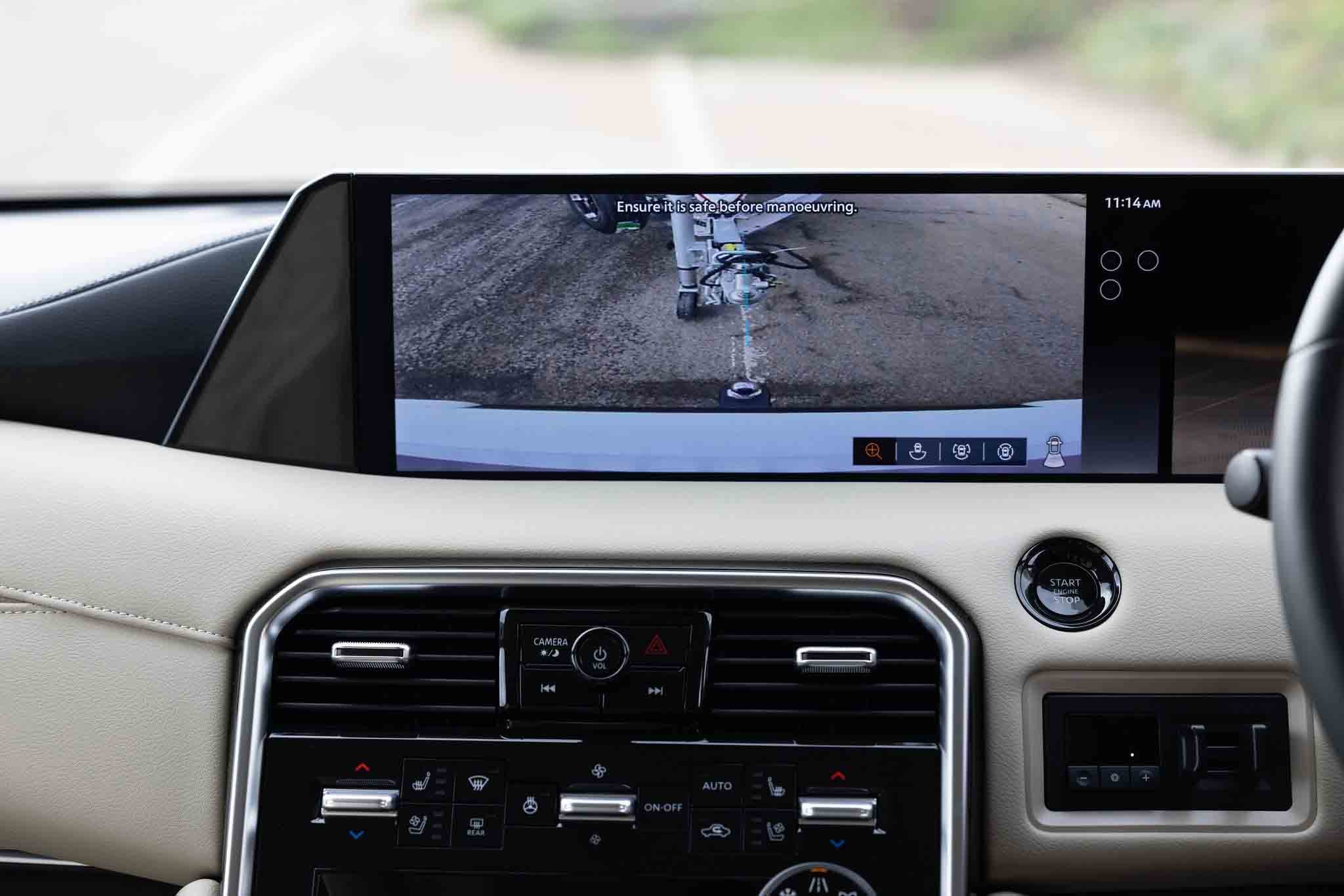 In-car dashboard screen showing the rear-view camera view with parking lines and the warning text 'Ensure it is safe before manoeuvring.'; clock reads 11:14 AM, car interior visible around the display.