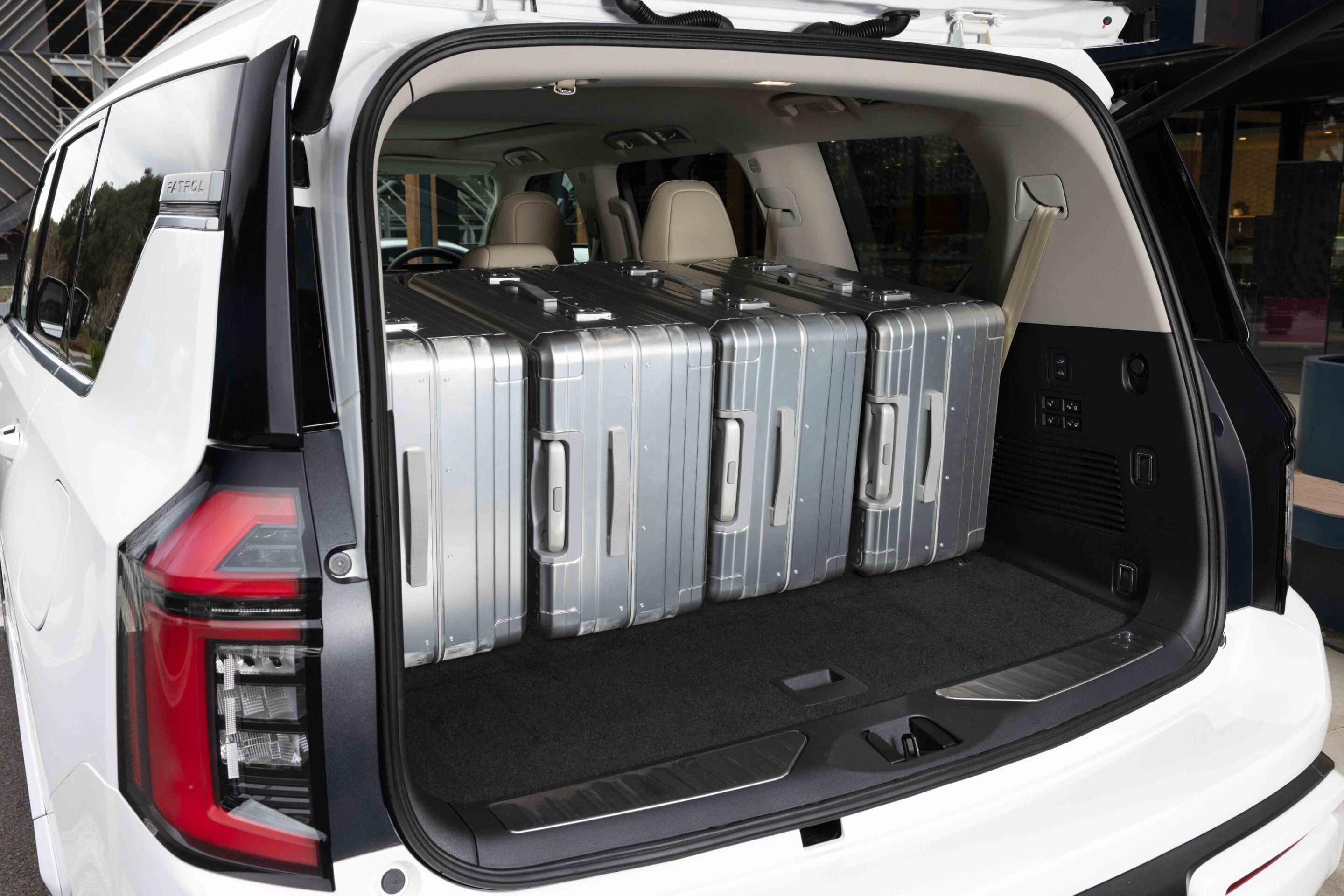 2026 Nissan Y63 Patrol finished in white, rear cargo area shown with the tailgate open, third-row seats folded flat and four large silver suitcases loaded inside.