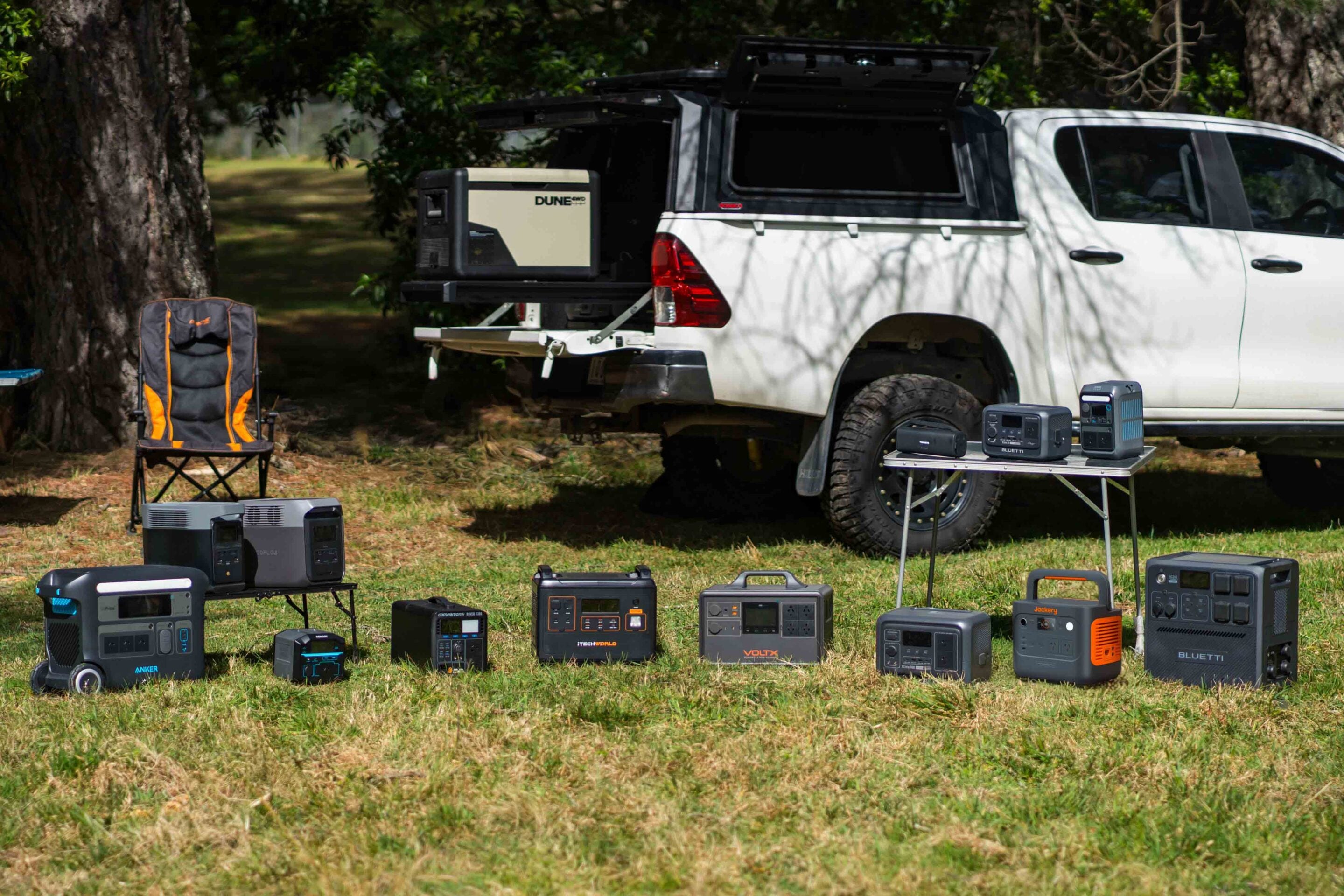White pickup truck with a camper shell parked under trees, several portable power stations and chargers spread across the grass and a table, suggesting an outdoor power setup.